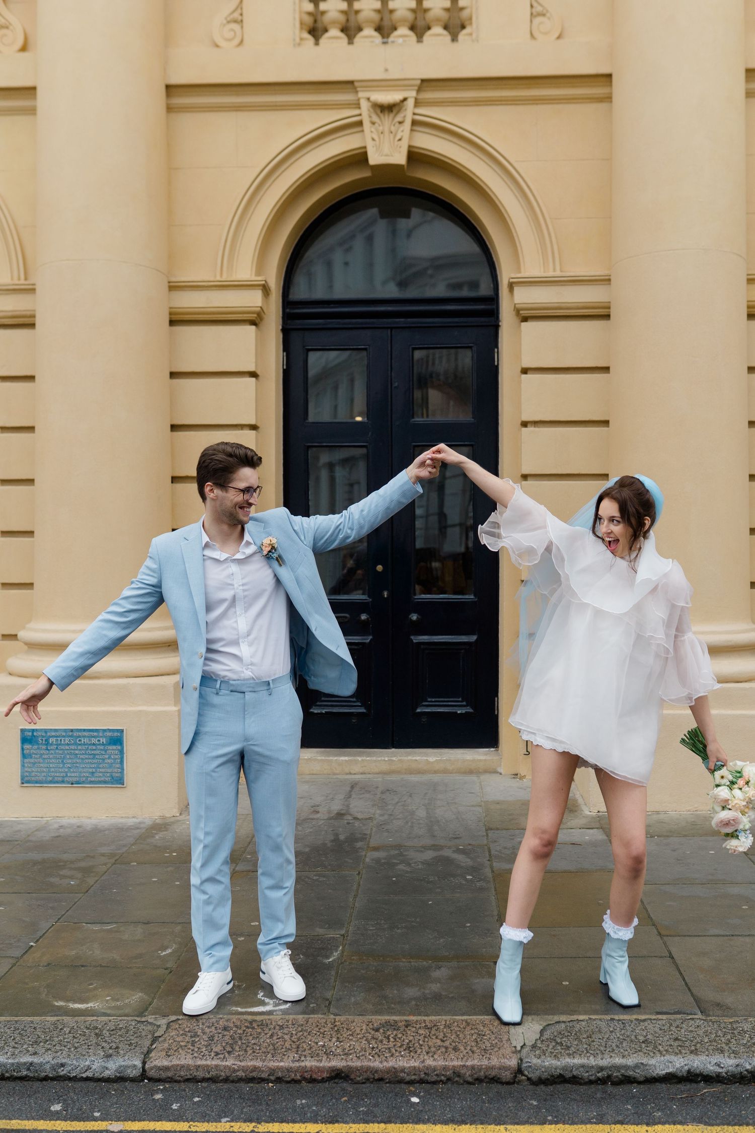 Couple in blue suit and white mini dress dancing outside elegant building on their wedding day.