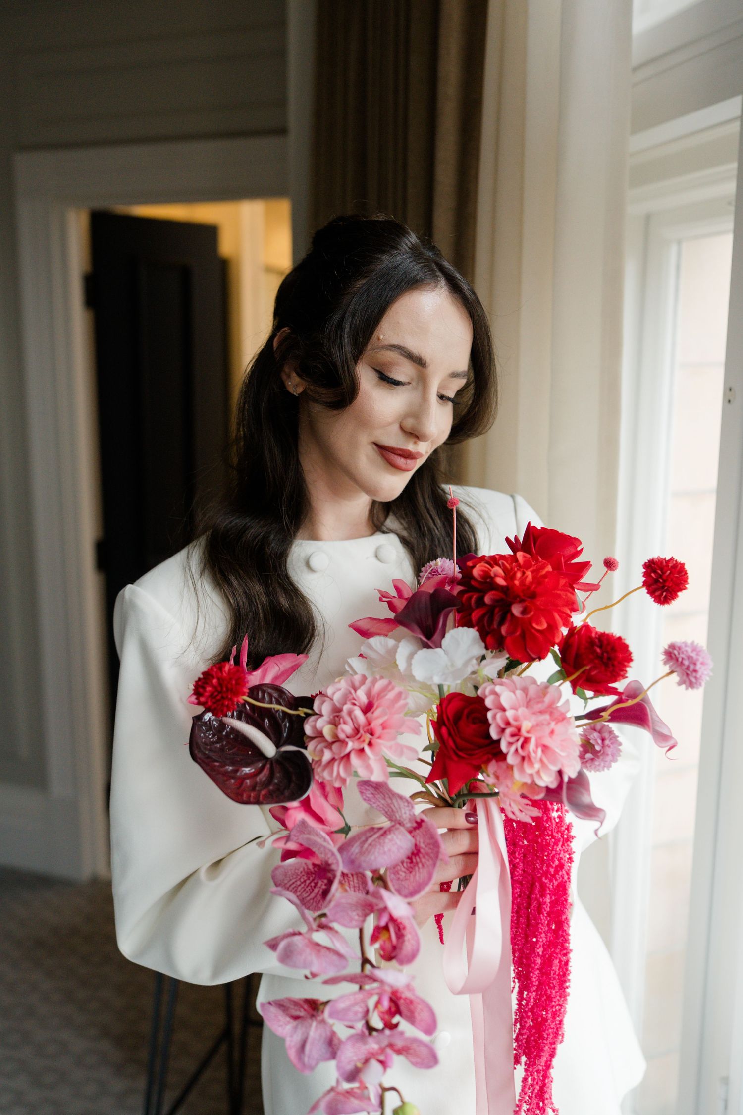 Elegant dark-haired bride in white dress gazes at stunning red and pink wedding bouquet by window.