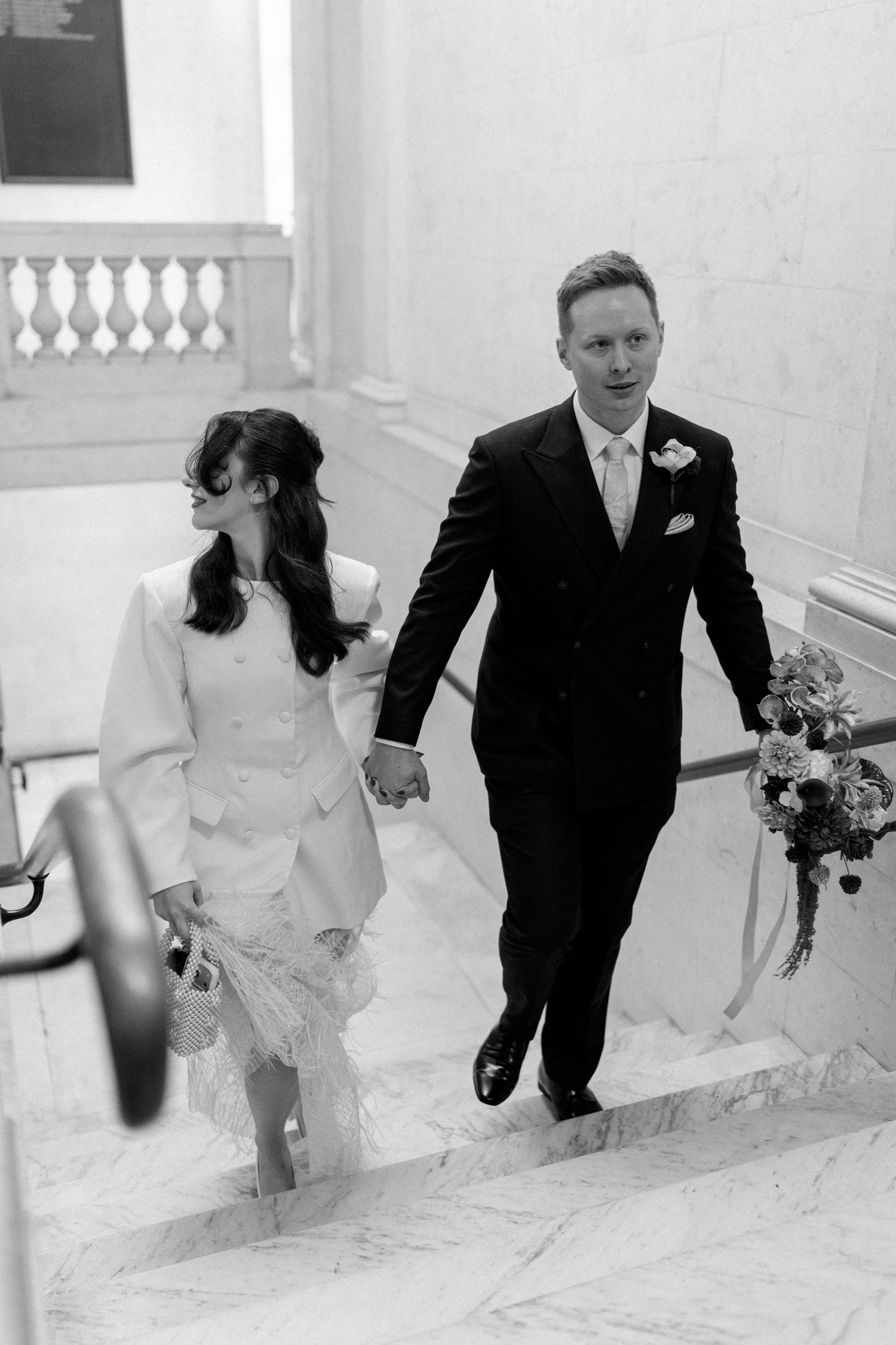 Black and white photo of newlyweds holding hands descending marble staircase after ceremony.