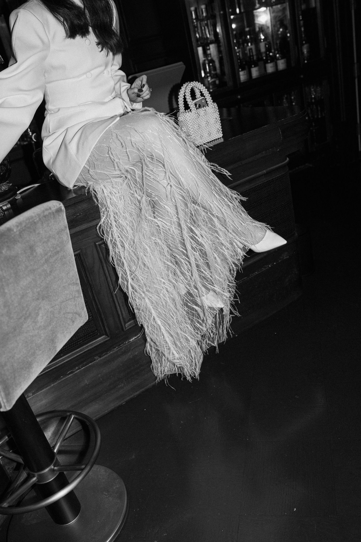 Black and white close-up of bride in feather skirt sitting at bar with pearl handbag.
