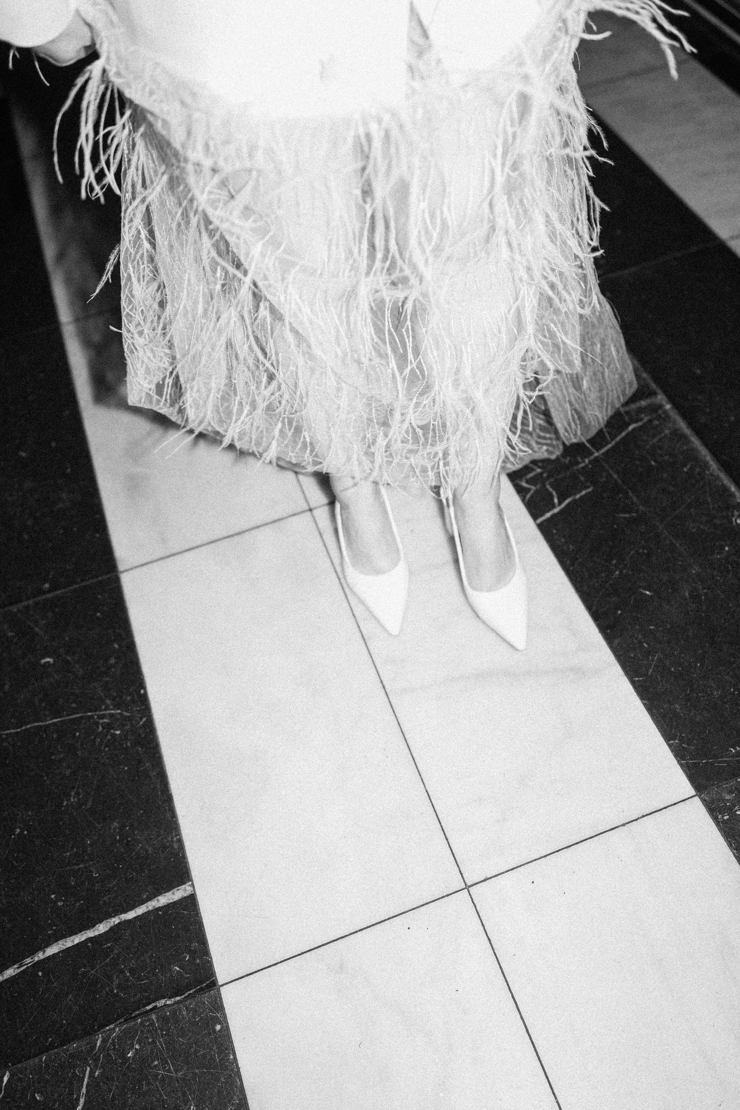 Close-up of bride's white pointed heels and feathered wedding skirt on black and white tiled floor.