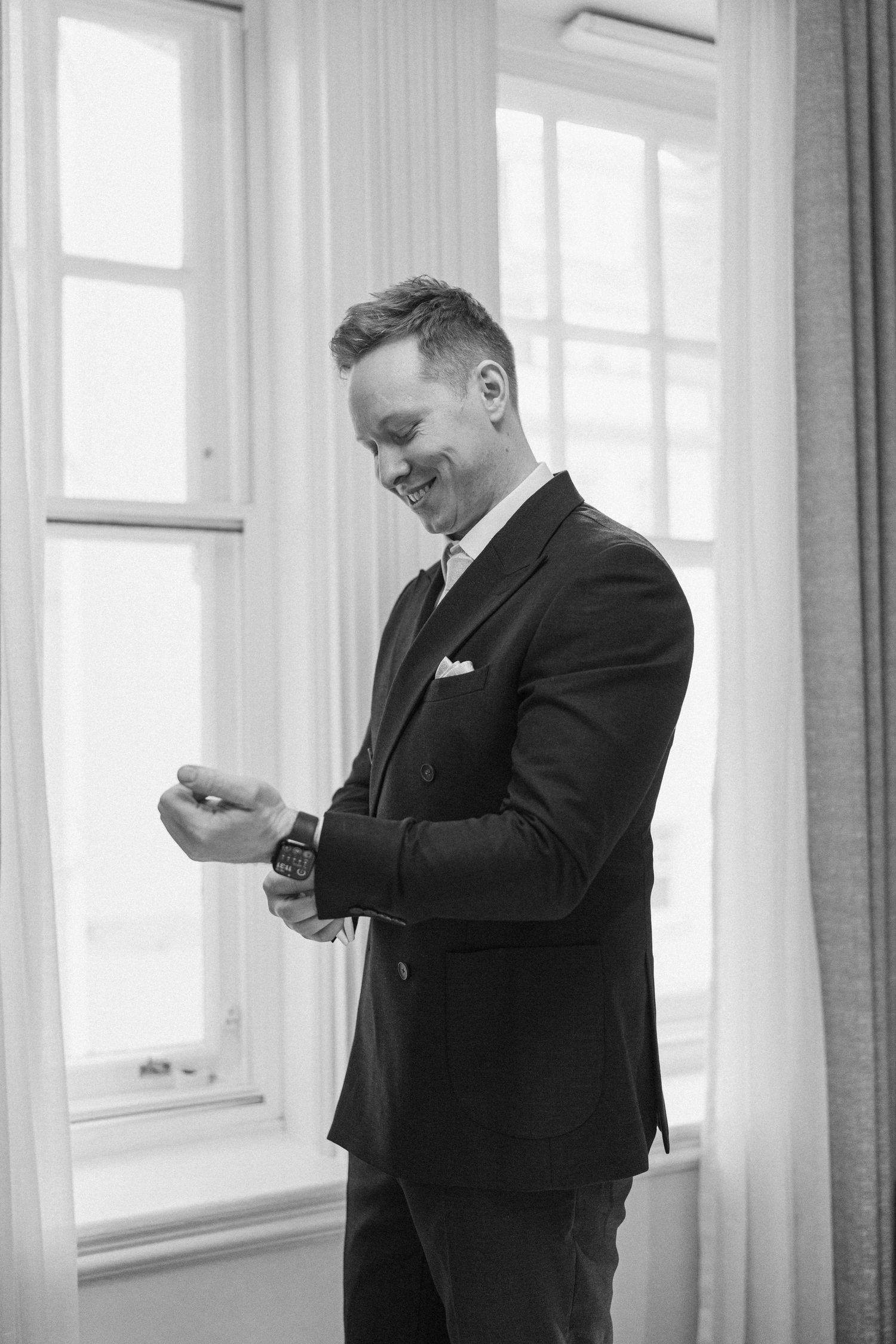 A smiling groom in a dark suit adjusts his cufflinks near bright windows in a black and white wedding photo.