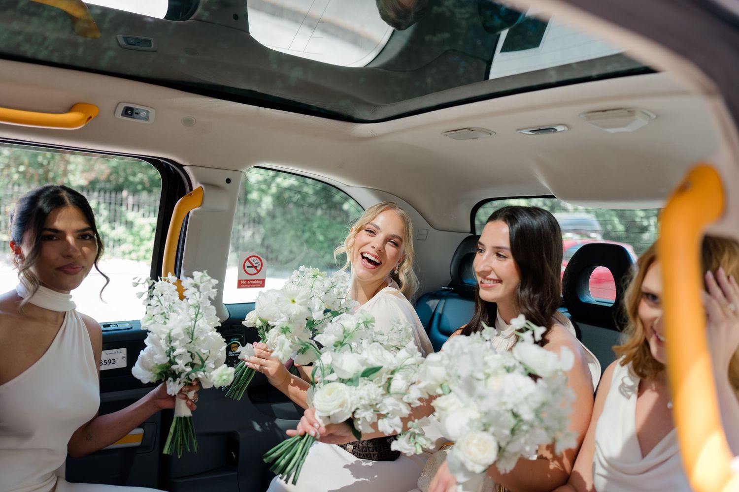 Laughing bride and bridesmaids in white dresses holding white floral bouquets inside a classic London black cab.