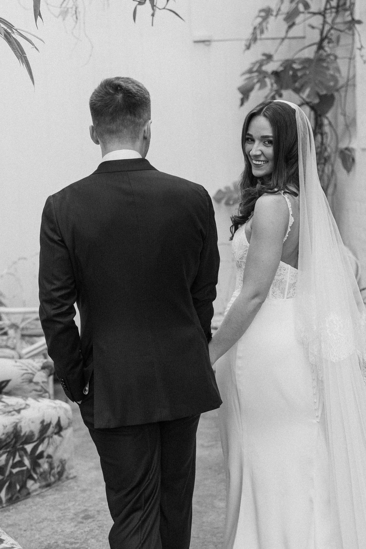 Black and white photo of smiling bride in lace wedding gown and veil holding grooms hand outdoors.