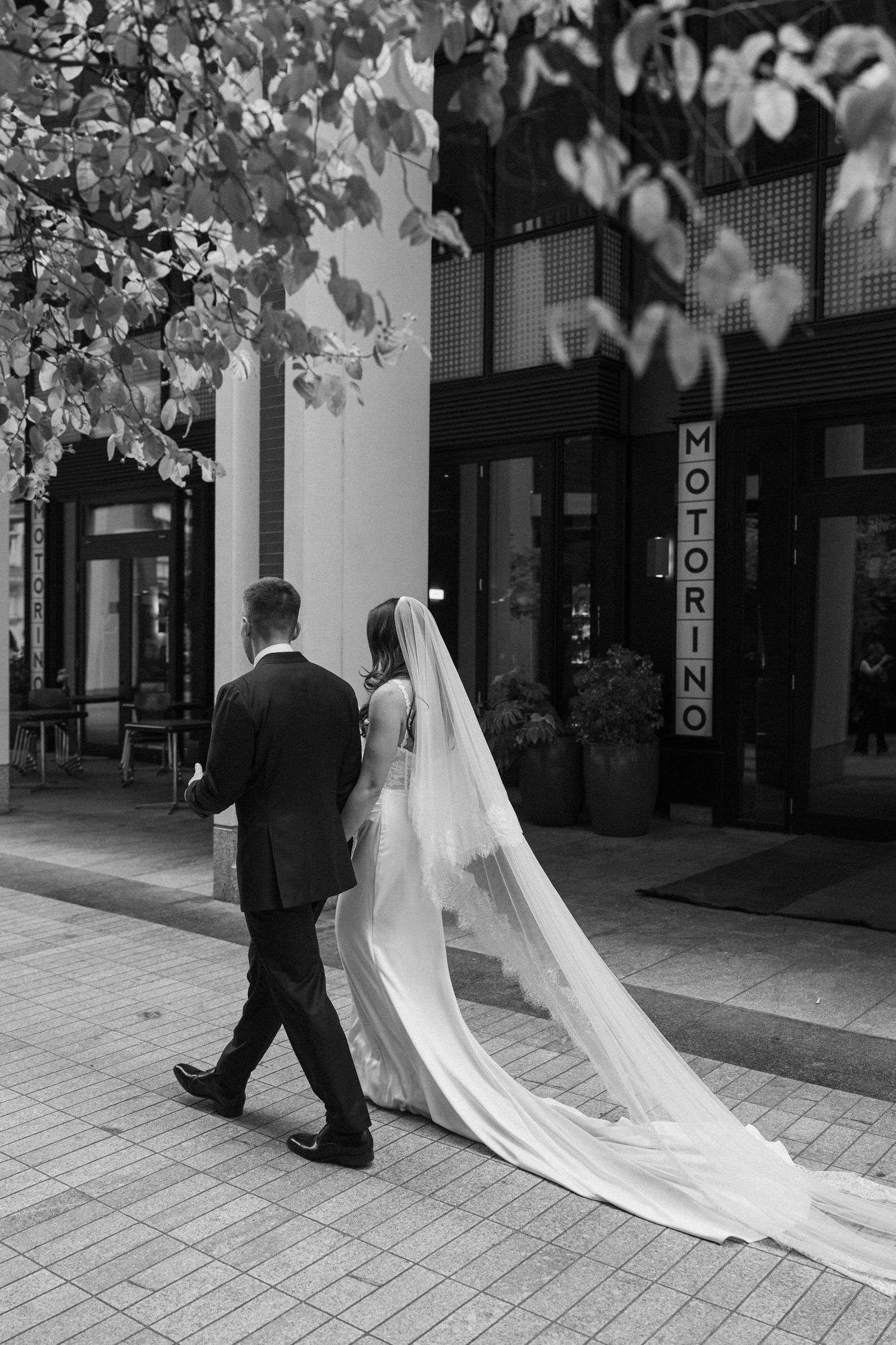 Black and white photo of bride and groom walking past a restaurant, bride's long cathedral veil trailing behind.