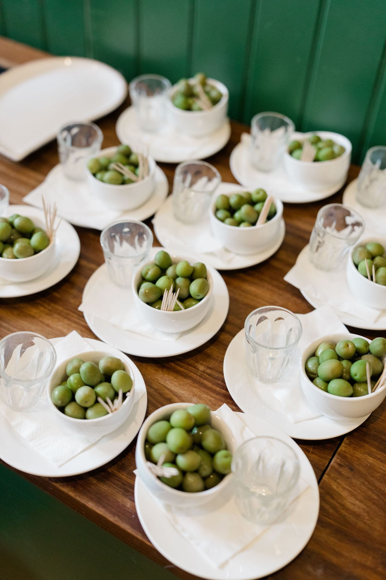 White bowls filled with green olives and toothpicks arranged on saucers with small glasses on a wooden table.