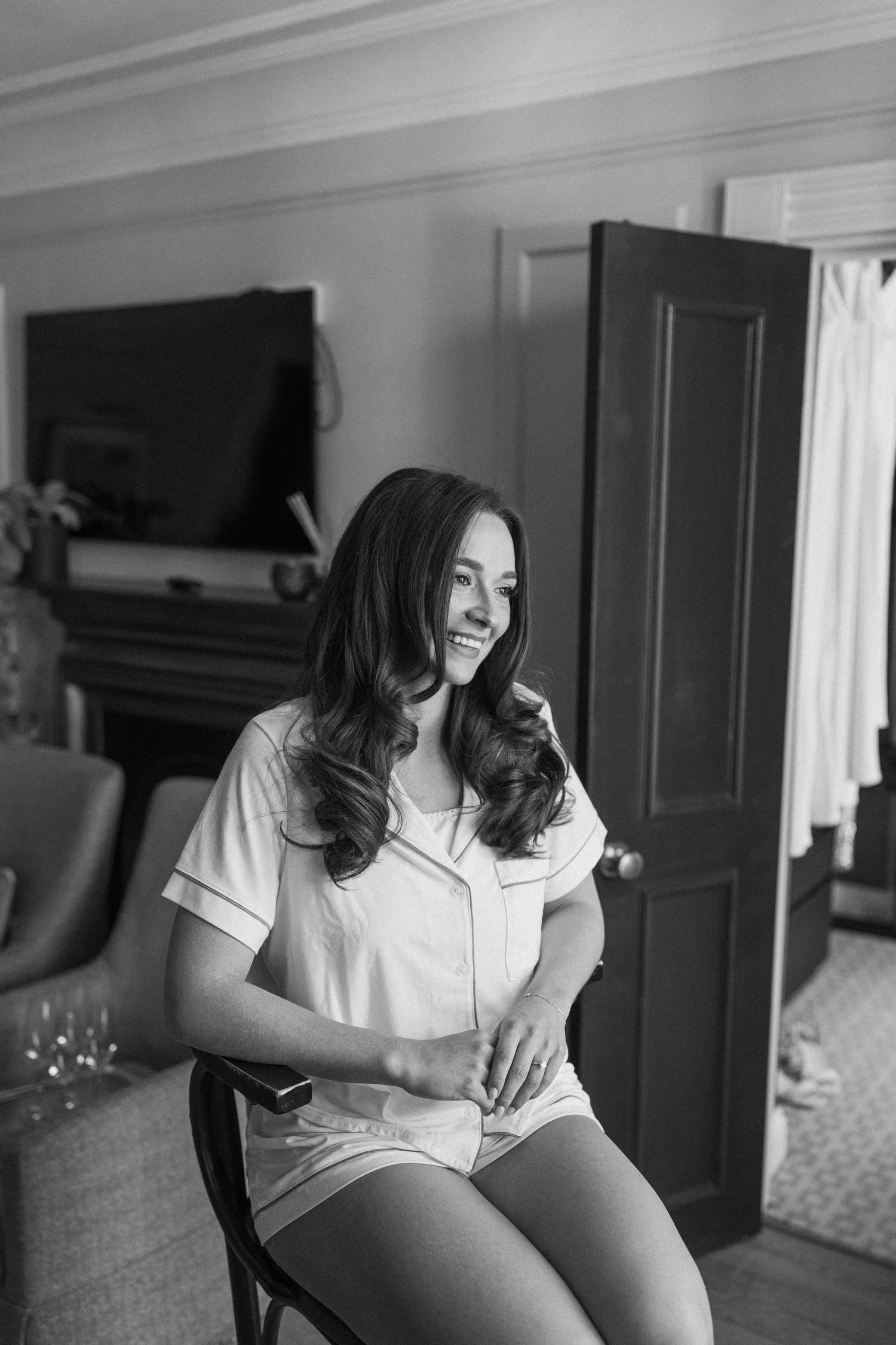 Smiling bride in white pajamas sitting in a chair during black and white wedding morning preparations.