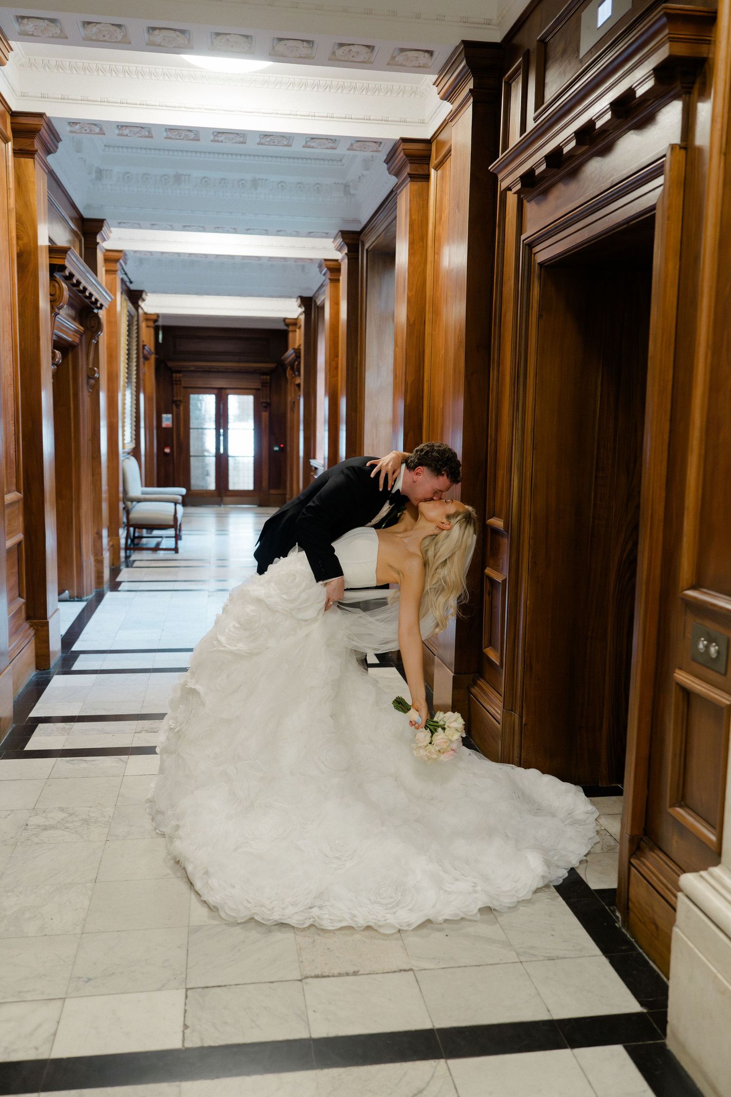 Bride and groom share a romantic dip kiss in an elegant wood-paneled hallway, bride in a flowing white gown.