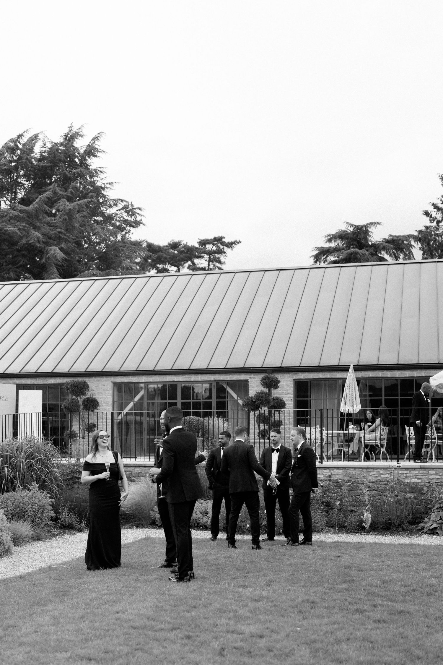 Black and white photo of wedding guests in formal attire socializing outside venue.