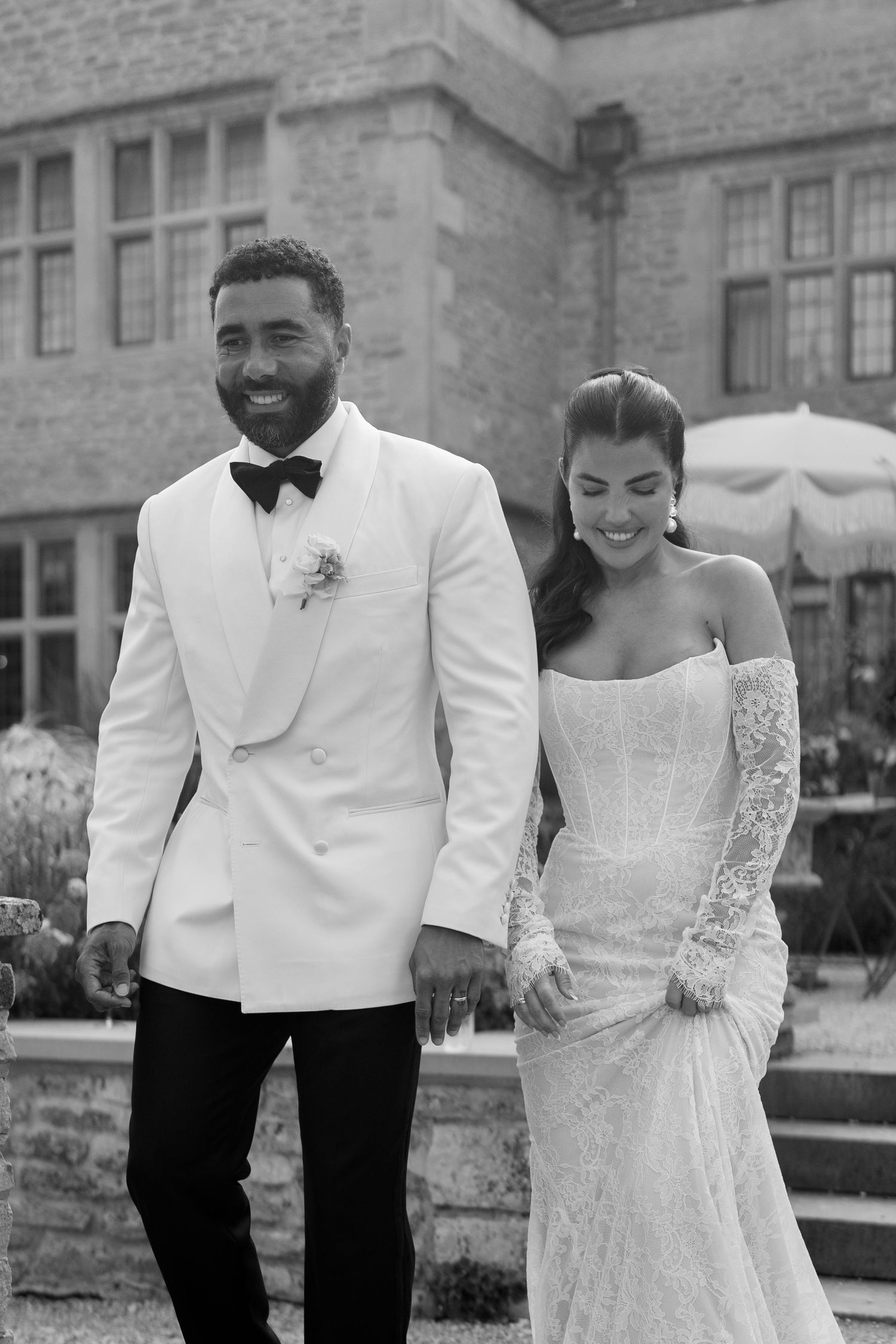 Black and white portrait of smiling bride and groom in white tuxedo outside a stone manor wedding venue.