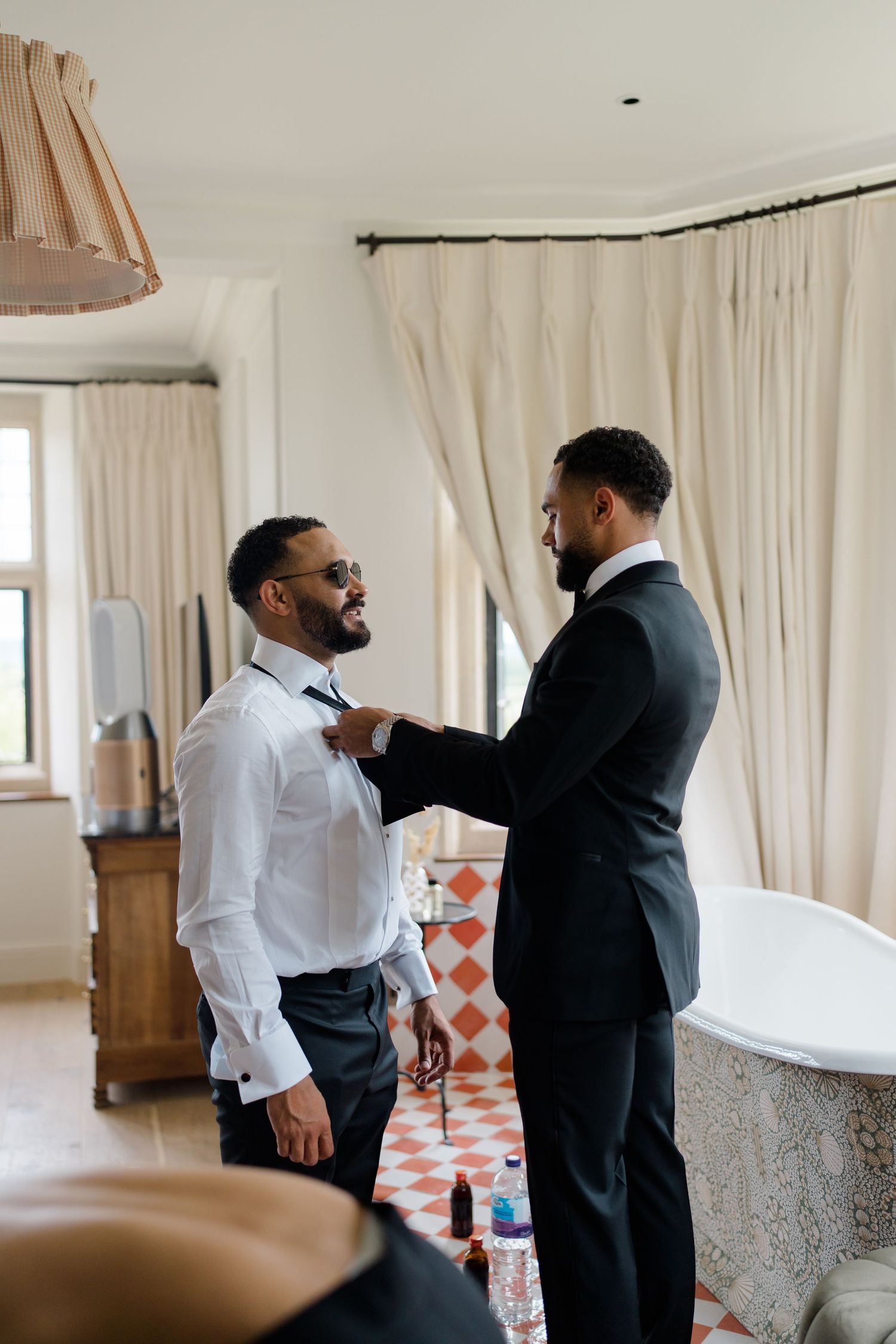 Two well-dressed groomsmen in black suits help each other with a bow tie in an elegant room.
