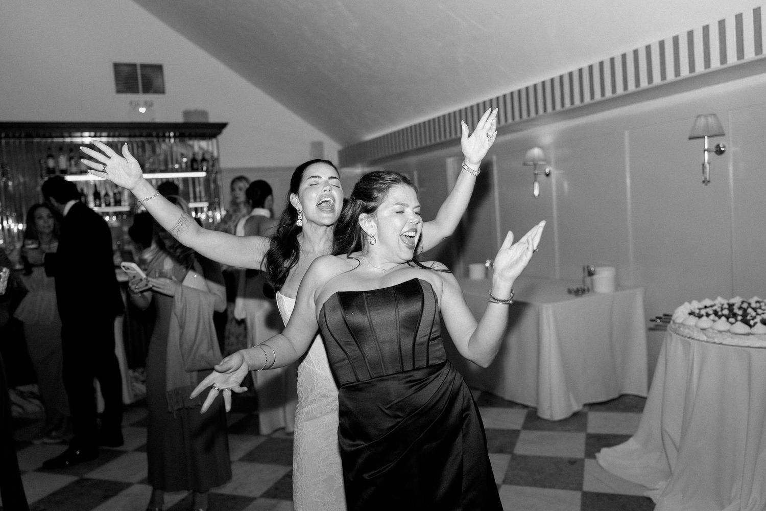 Two women laugh and dance energetically on a black and white checkered wedding reception dance floor.