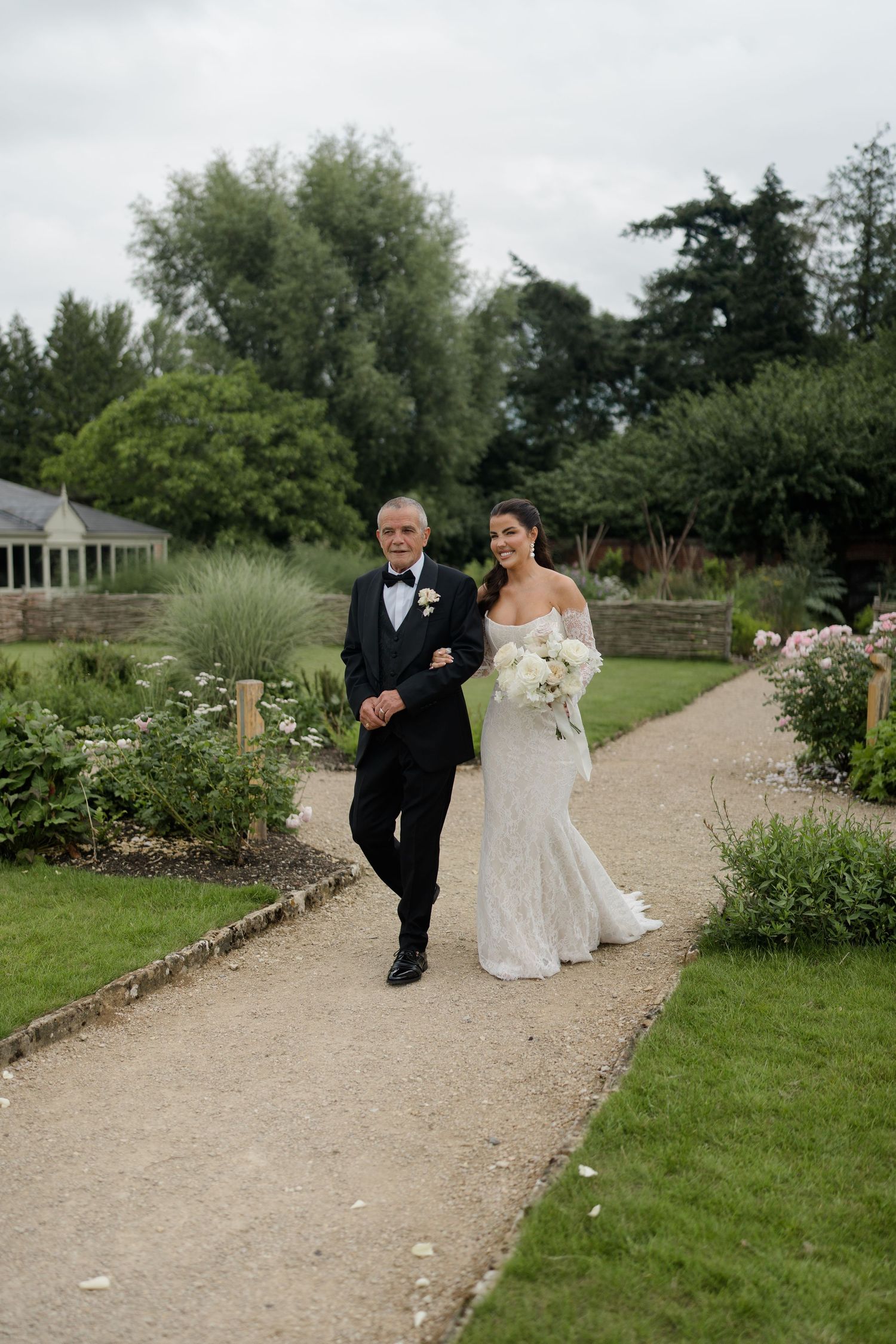 A bride in a white gown walks with her father down a garden path on her wedding day.