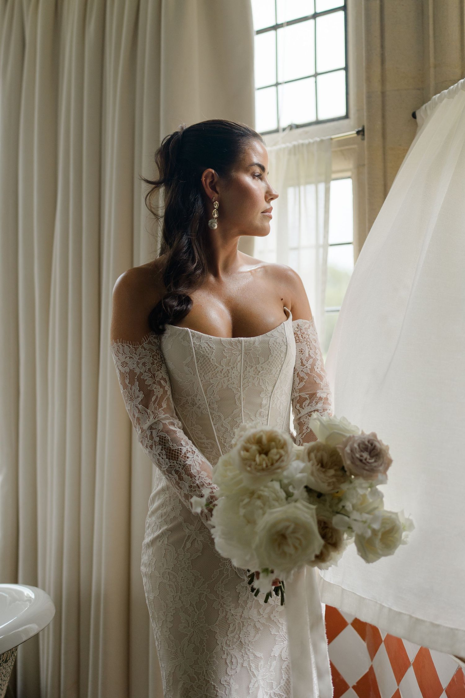 Bride in an off-shoulder lace wedding gown holding a bouquet of white roses by a window.
