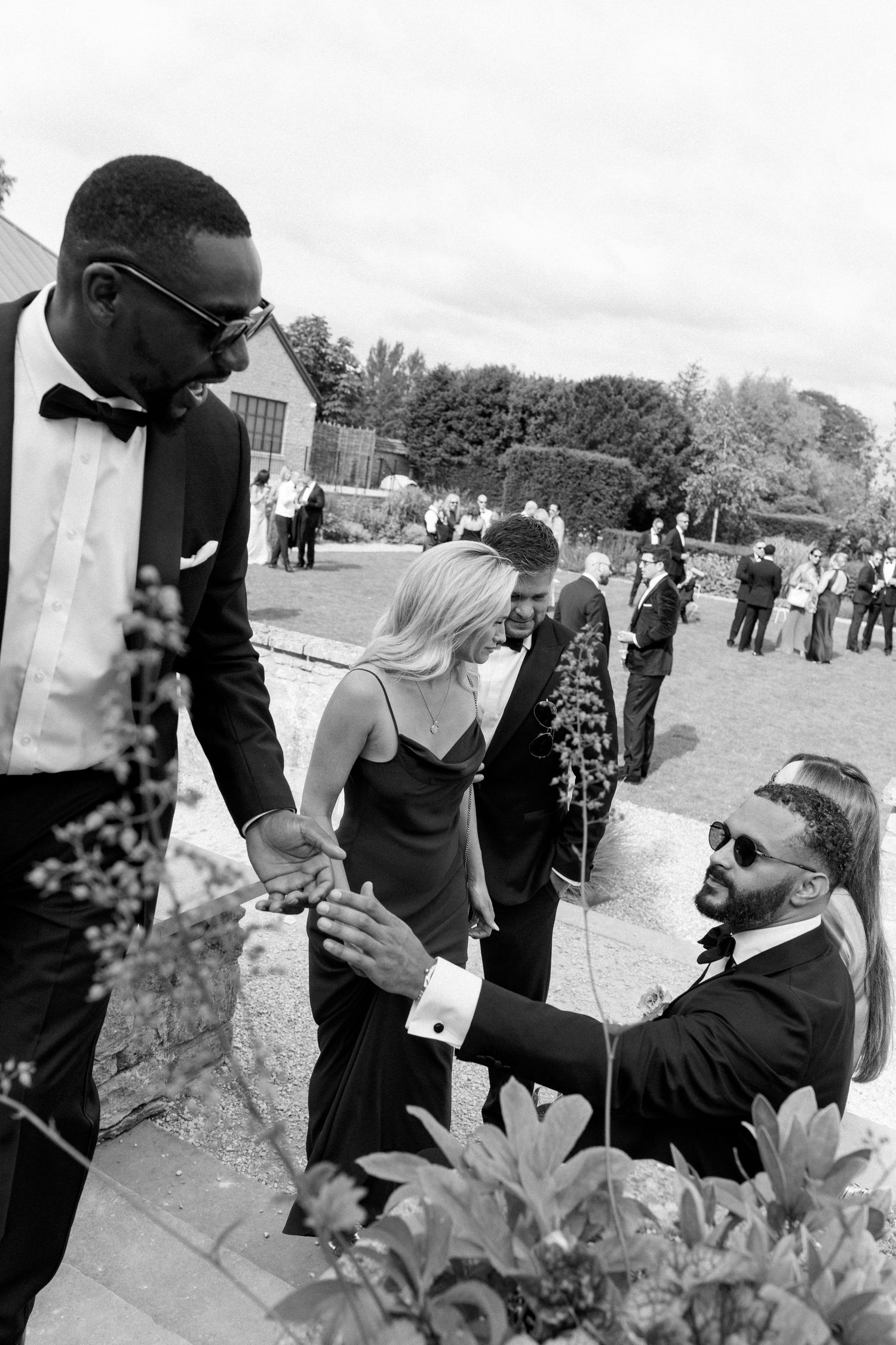 Black and white photo of smartly dressed wedding guests greeting each other warmly in a garden setting.