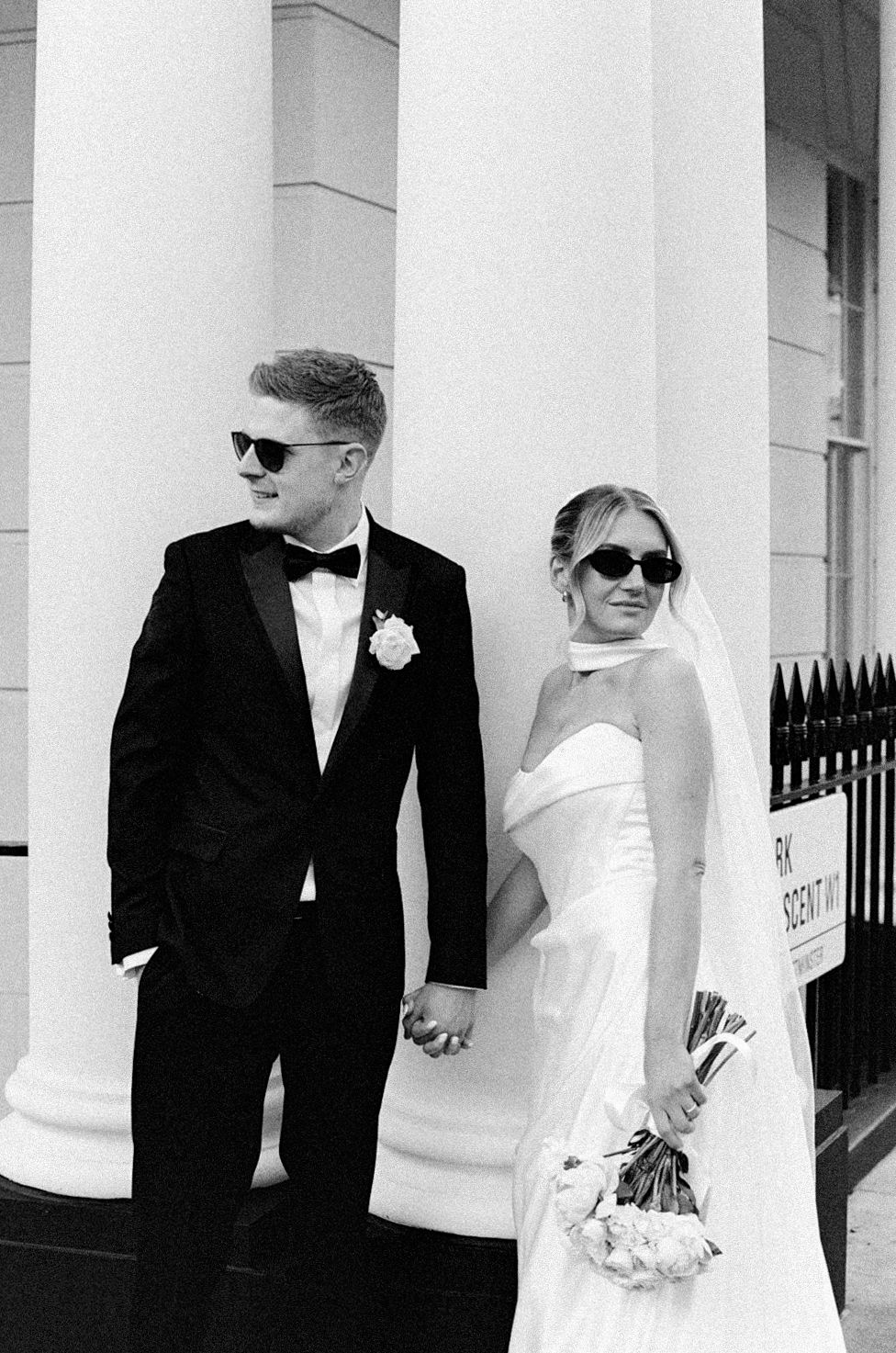 Black and white photo of a bride and groom holding hands in sunglasses, posing by white columns on their wedding day.