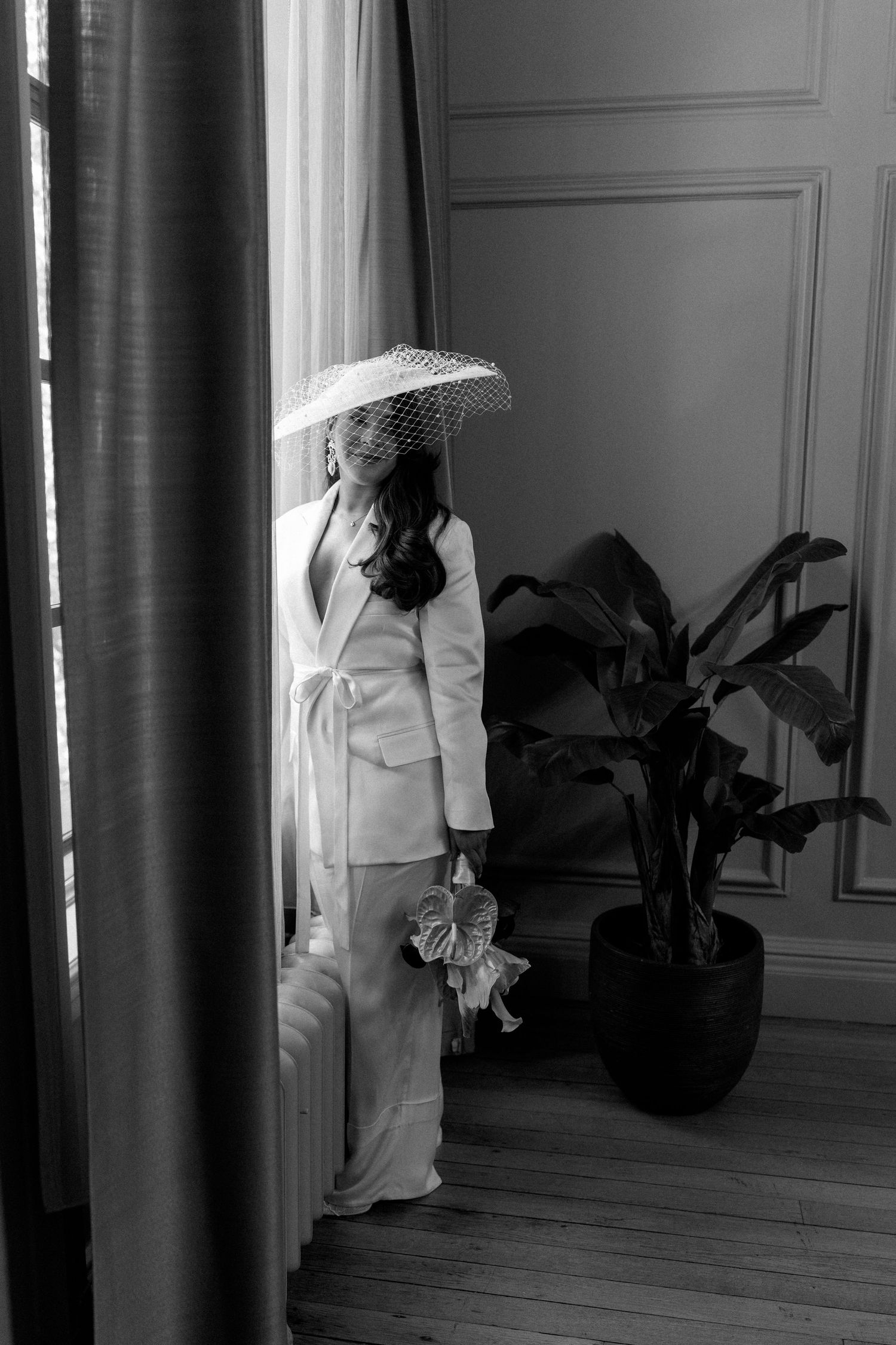 Black and white photo of a bride in a white suit and birdcage veil holding a bouquet by a window.