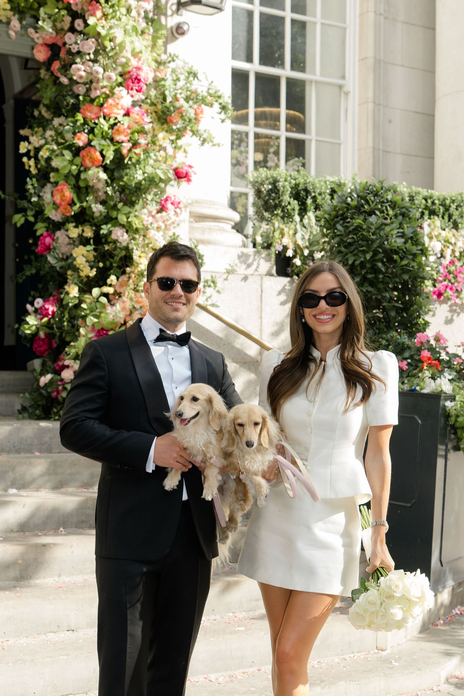 Couple in wedding attire with dachshund dog posing before floral-decorated building entrance.