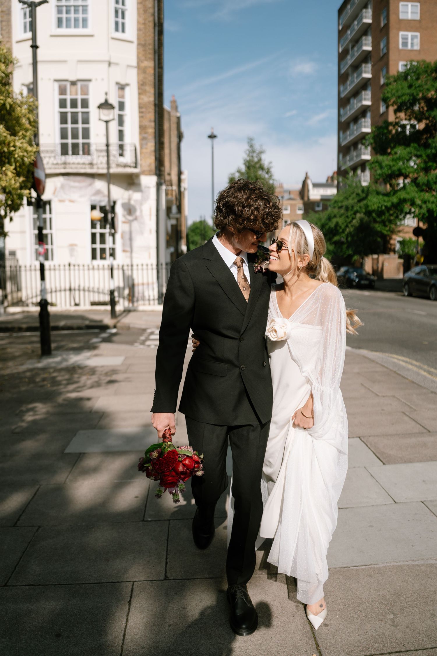 Stylish couple in wedding attire walking together on a sunny urban sidewalk.