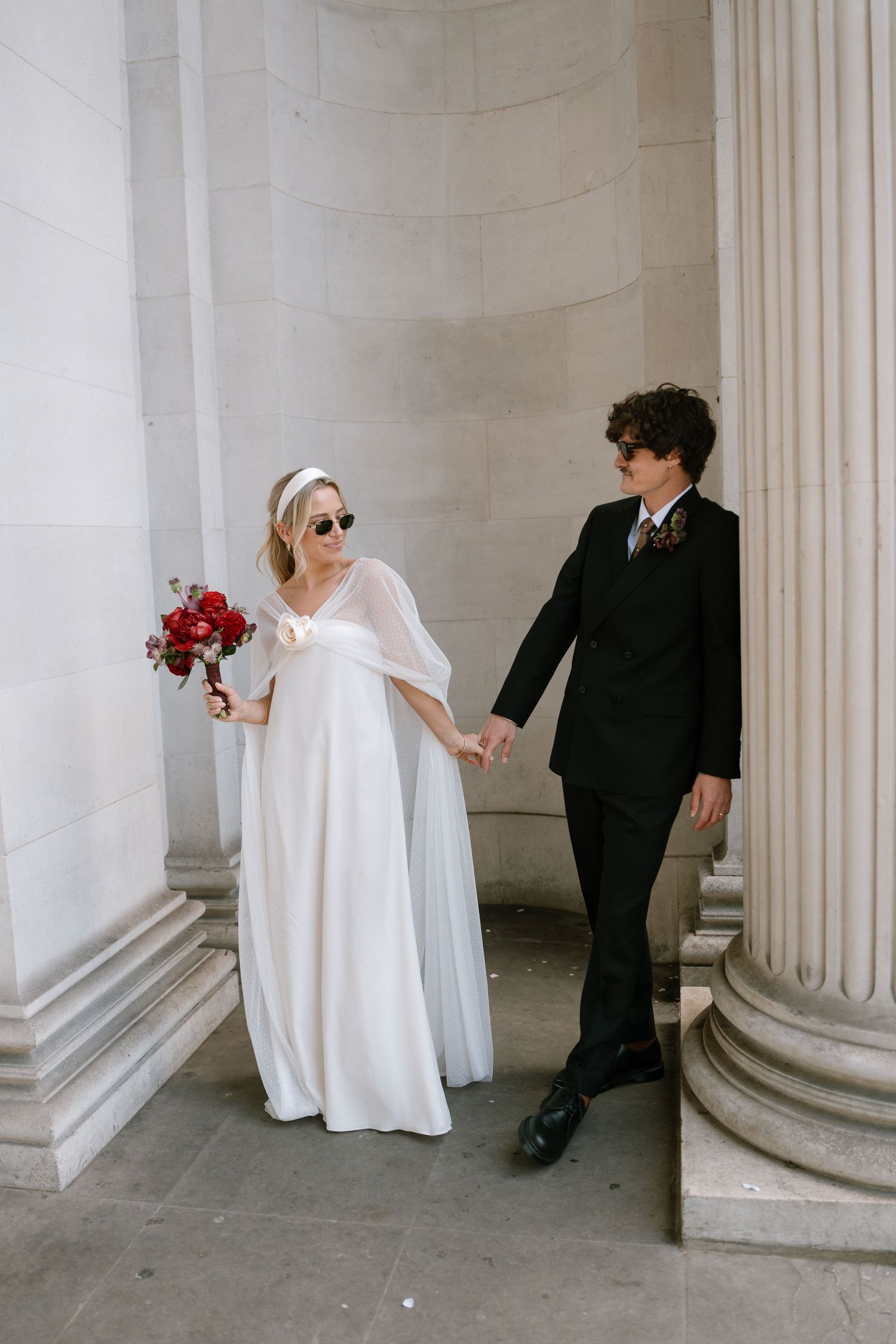 A bride in sunglasses holds red flowers while standing with groom between classical columns.