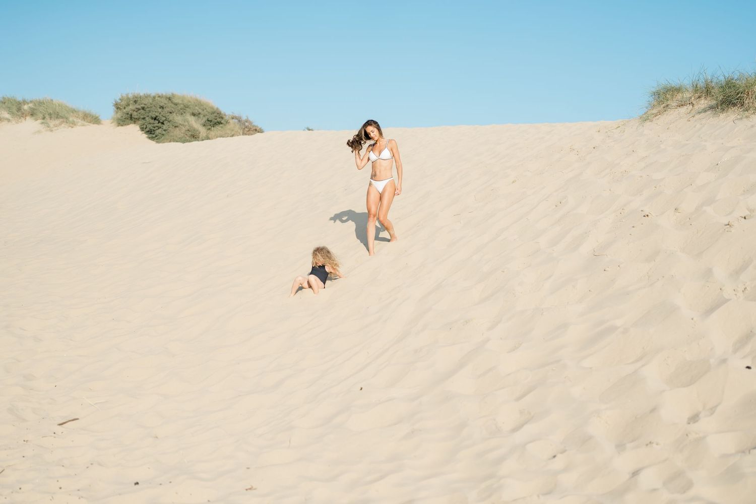Two figures descending a steep sand dune together on a bright sunny day.