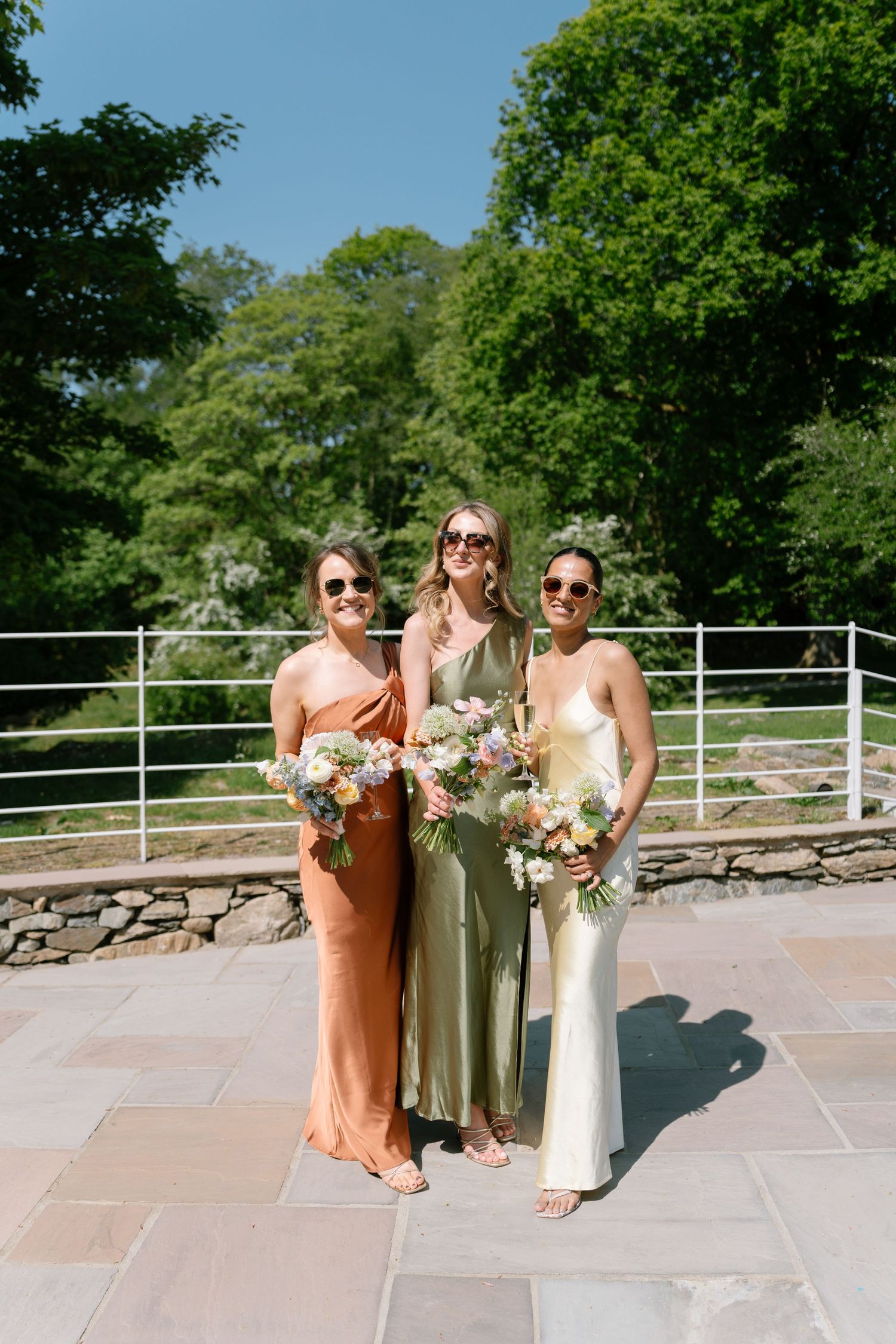 Bridesmaids in peach and cream dresses hold bouquets while standing on a terrace.