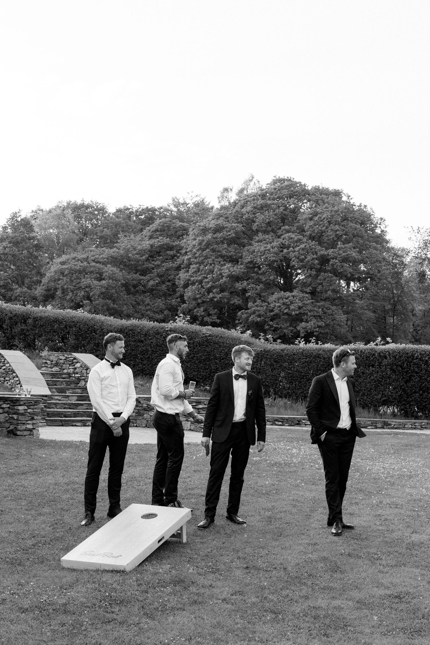 Groomsmen in black tuxedos and bowties stand in a row on grassy lawn.