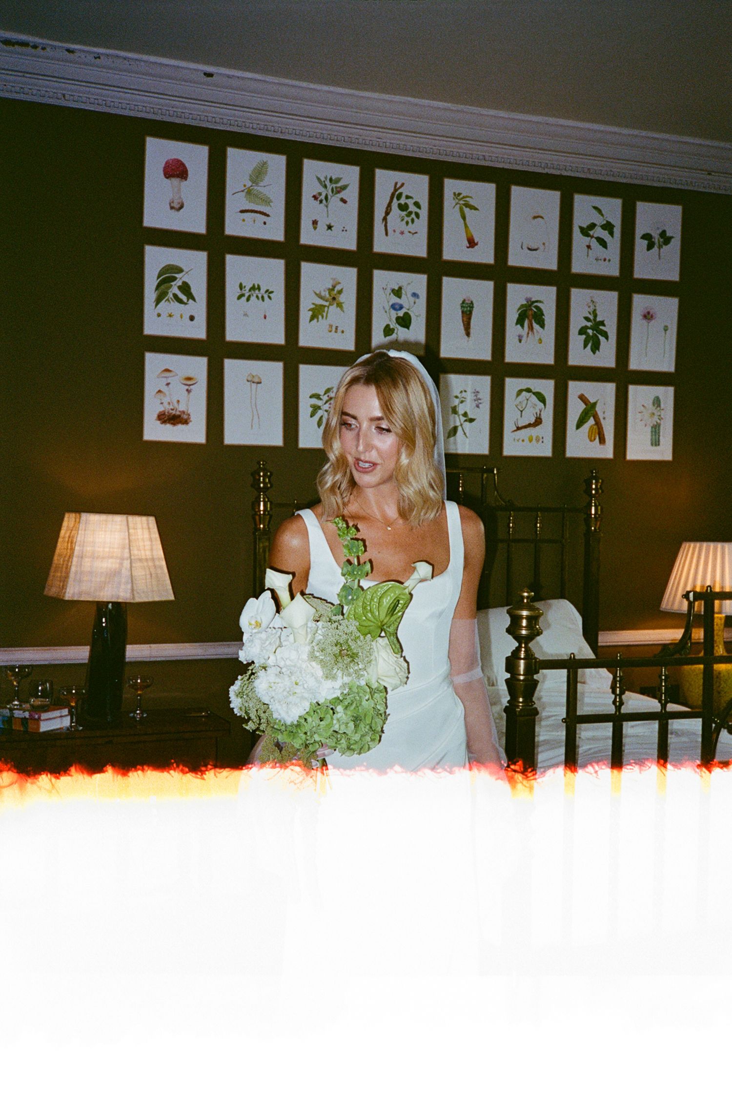 Blonde bride in white dress holds green and white bouquet in a room with botanical prints on dark walls.