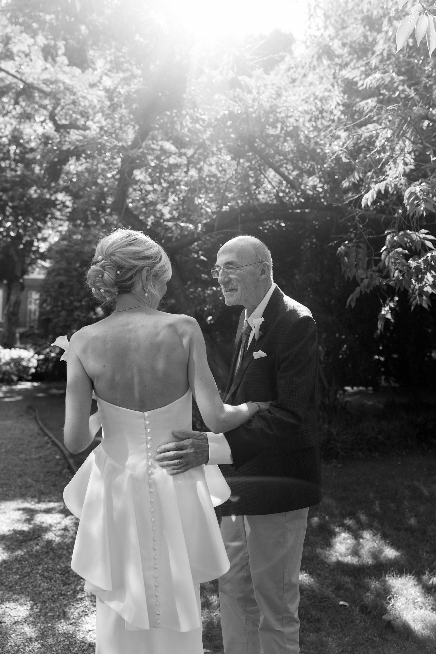 Black and white photo of a bride in a backless gown sharing a tender moment with an older man in a sunny garden.
