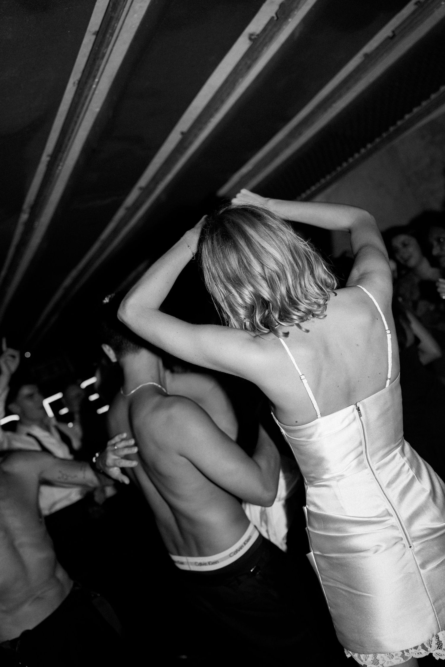 Bride in white slip dress dances with a shirtless guest on a crowded wedding dance floor.