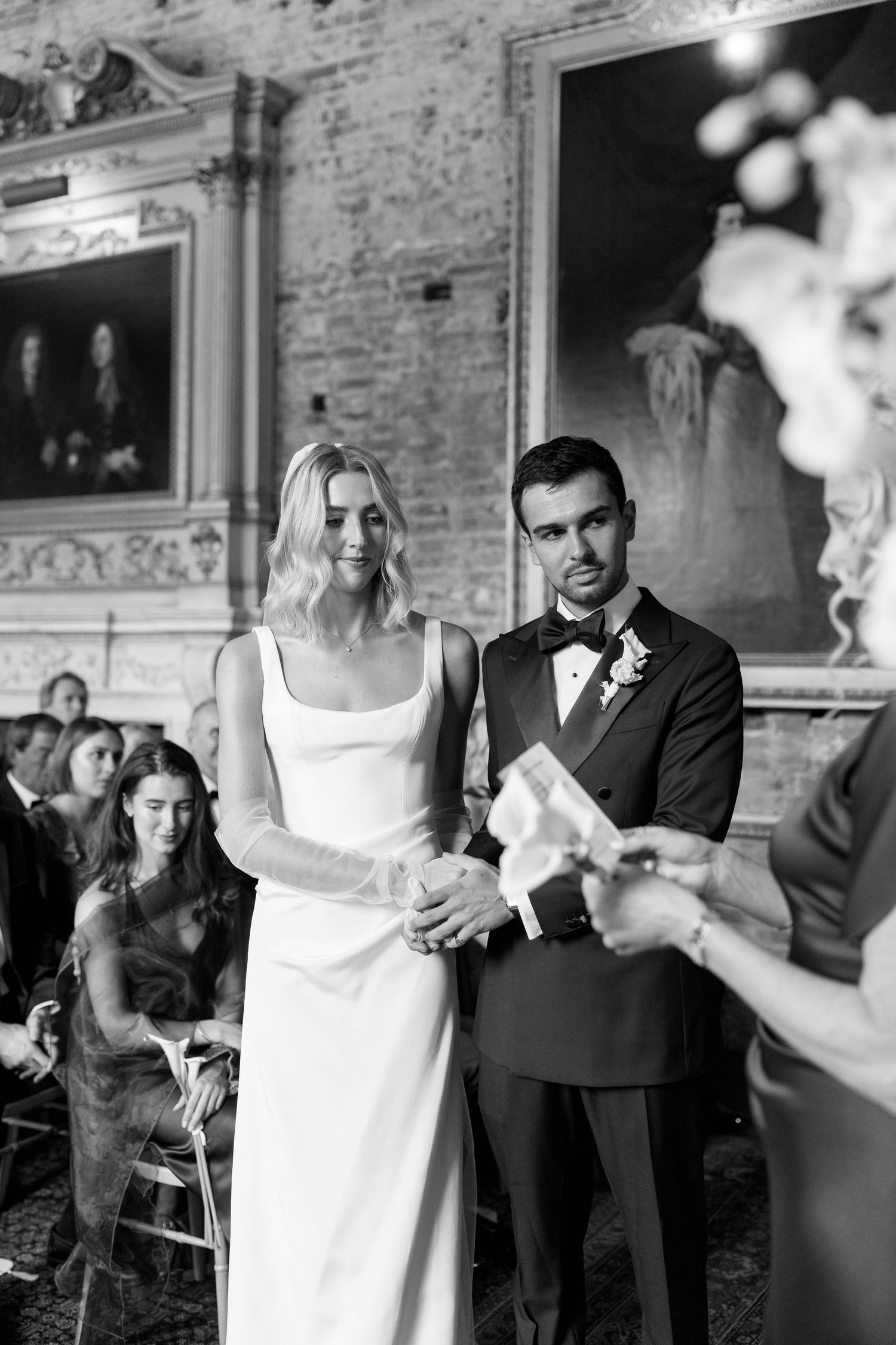 Black and white photo of bride and groom holding hands at wedding ceremony in elegant hall.