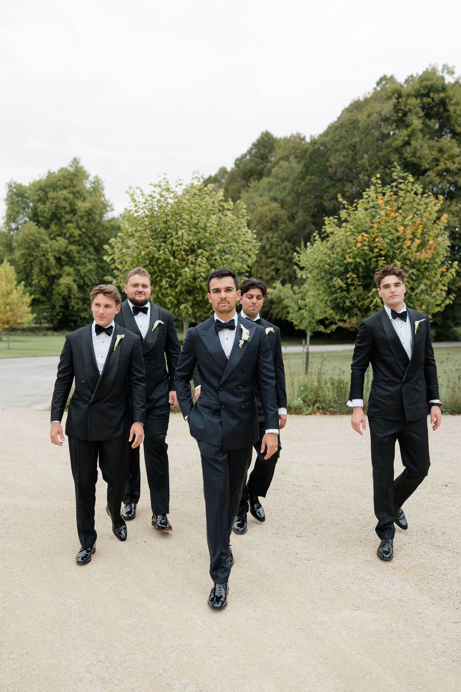 Groom and groomsmen in black tuxedos with bow ties walking confidently outdoors at a wedding venue.