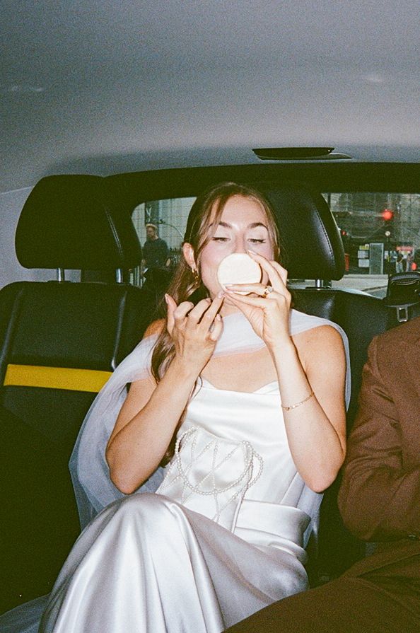A young woman in a white satin dress sits in a car's back seat, holding a compact mirror to her face.