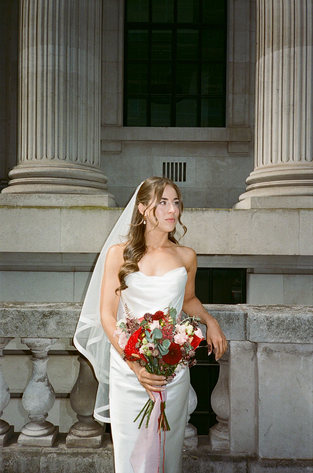Bride in white cowl-neck gown and veil holds red bouquet beside stone columns.
