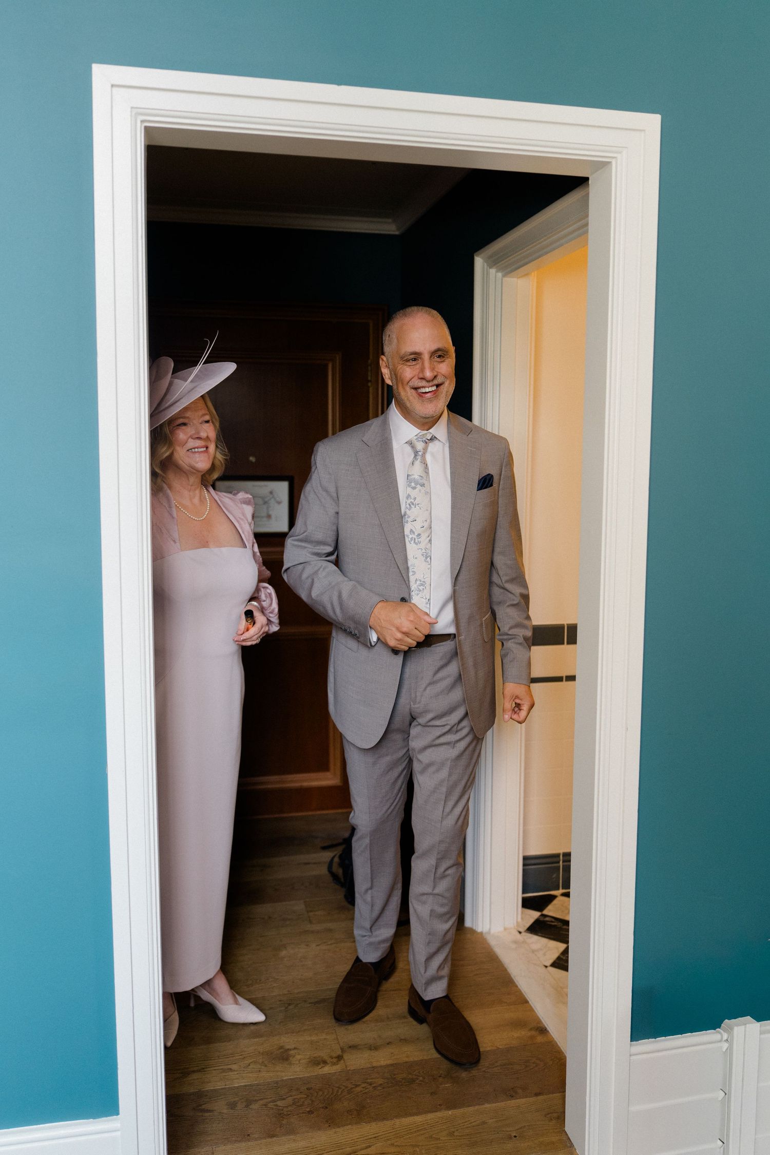 Smiling groom in grey suit stands in doorway with elegantly dressed woman in pink beside him.