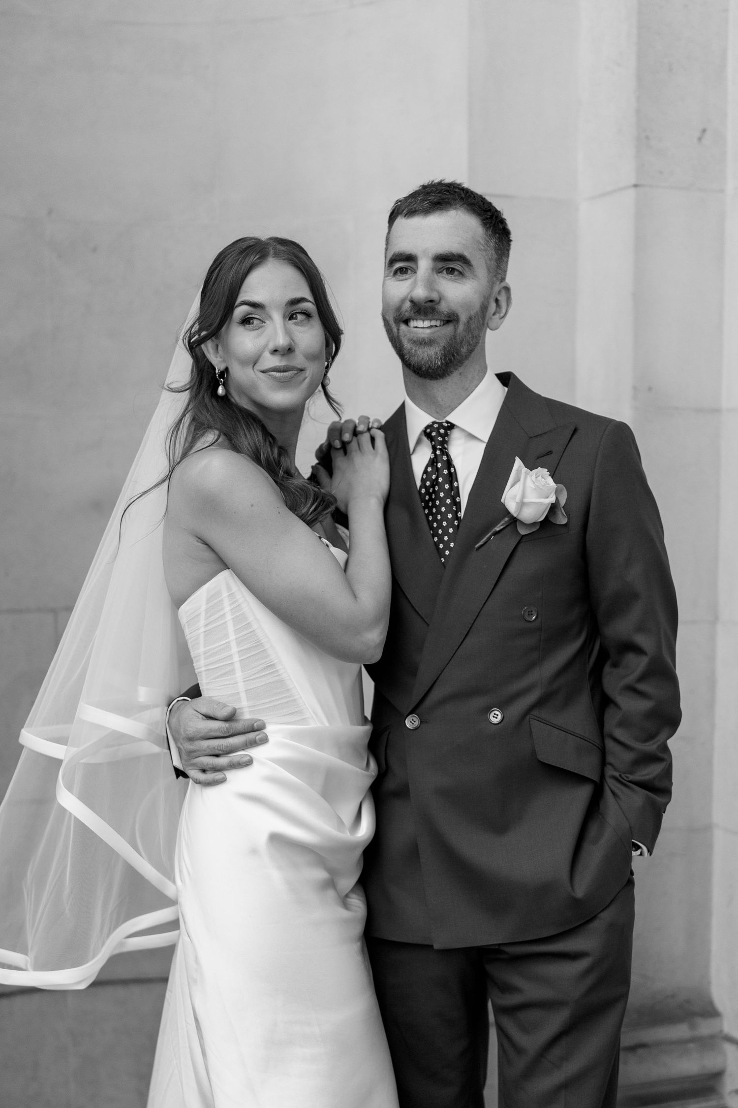 Black and white portrait of smiling bride and groom embracing outside elegant stone building on wedding day.