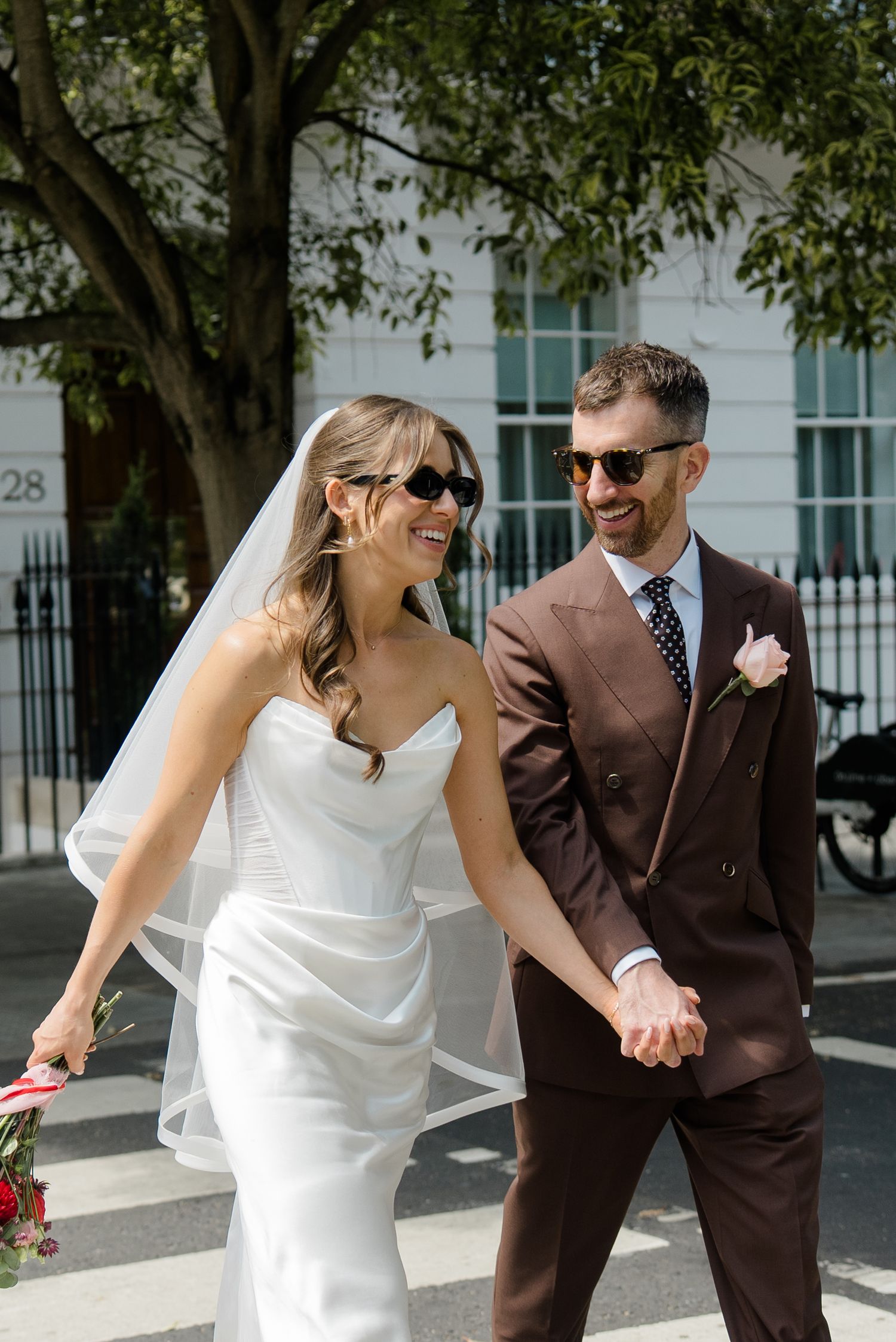 Smiling bride and groom in sunglasses hold hands while crossing a sunny London street.