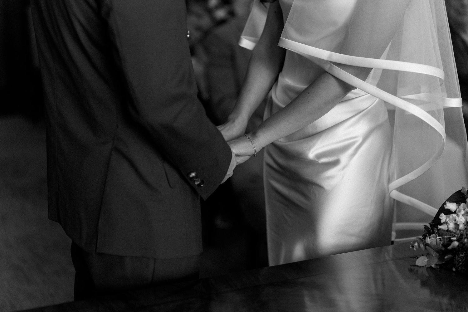 Black and white close-up of bride and groom holding hands at their wedding ceremony altar.
