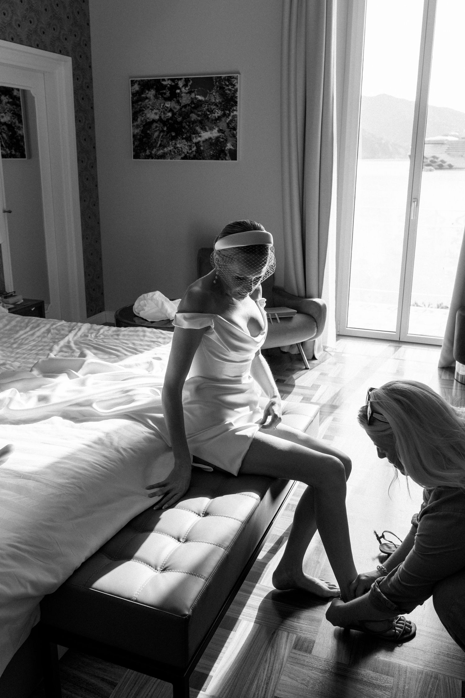 Bride in white dress sits on bed bench while assistant helps with shoes in elegant hotel room.