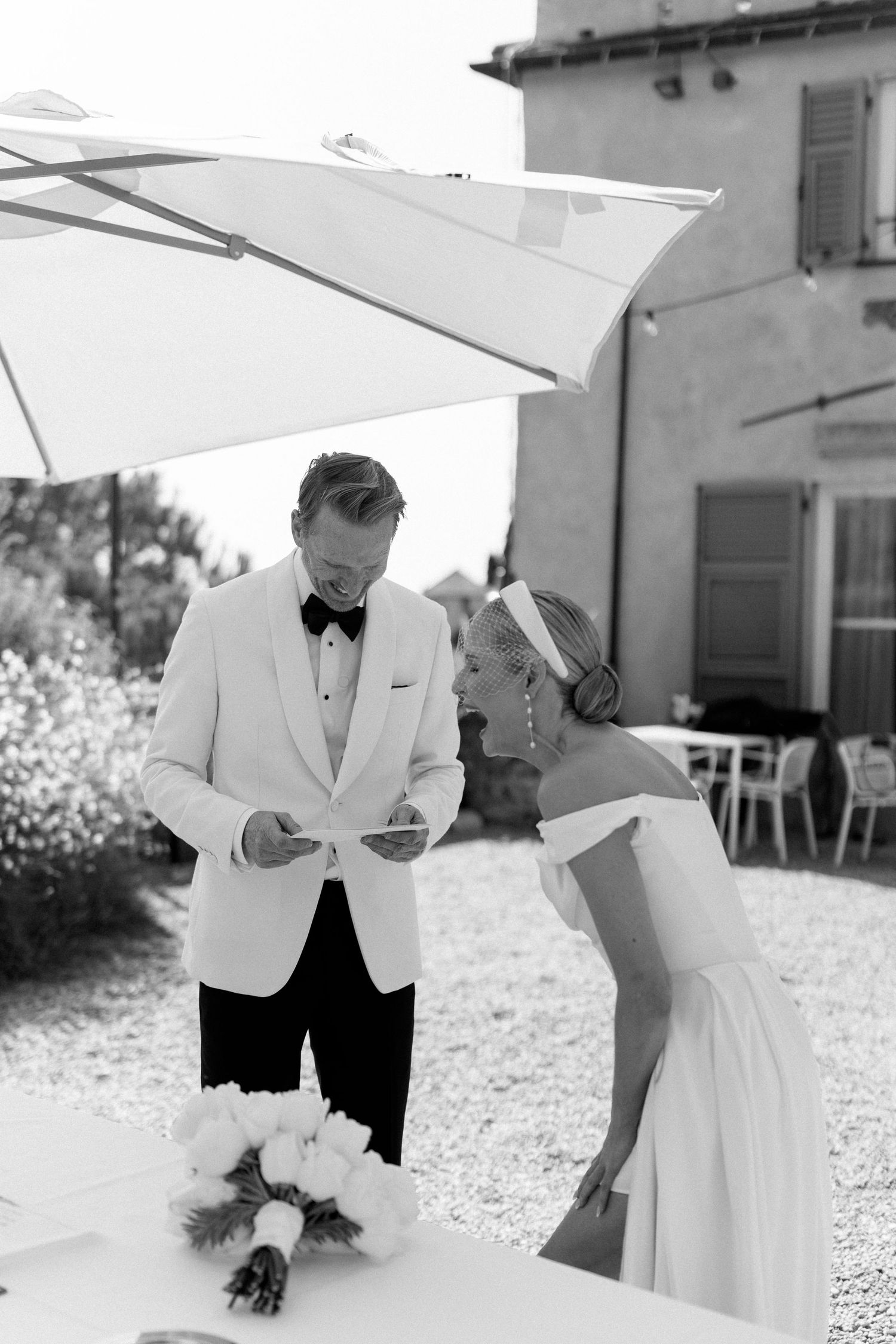 Black and white photo of bride and groom reviewing wedding documents under an umbrella at an outdoor Italian venue.