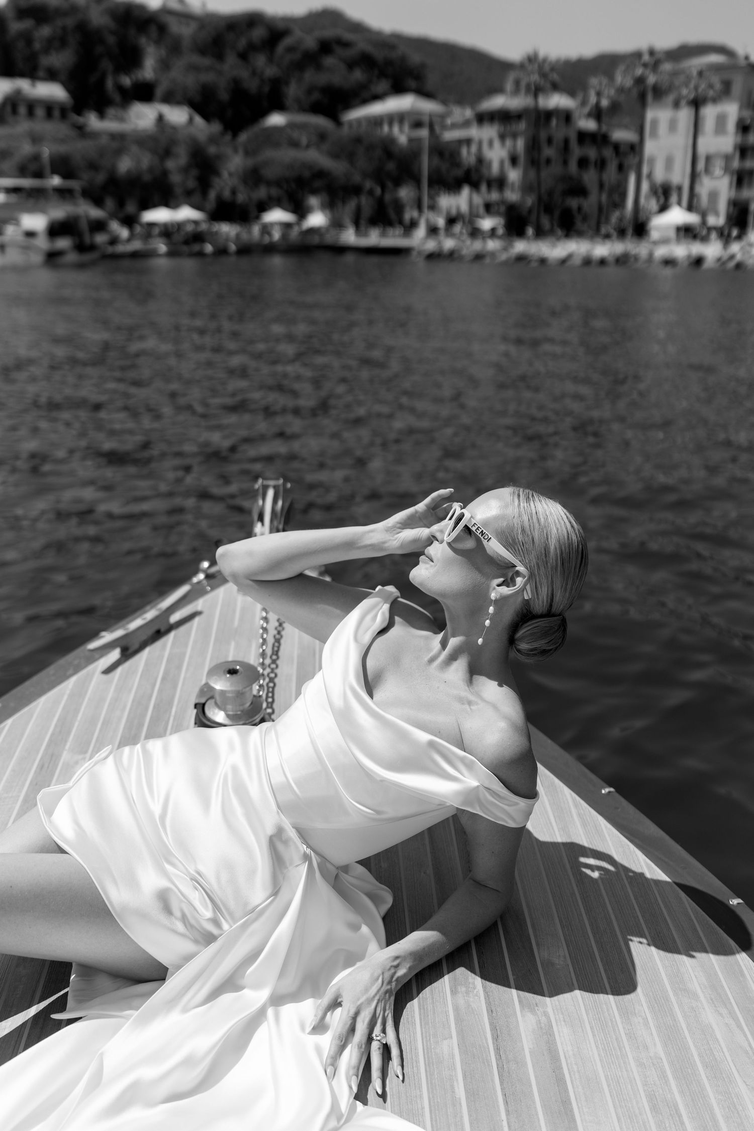 Glamorous bride in white dress reclines on wooden boat deck holding sunglasses with coastal town in background.