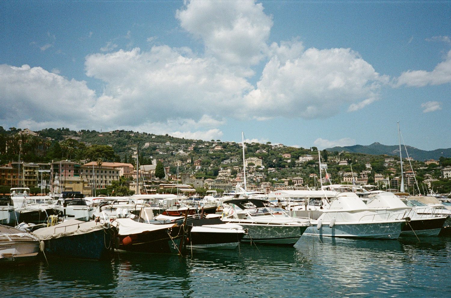 Luxury yachts and motorboats moored in a bustling Italian Riviera marina under a partly cloudy sky.