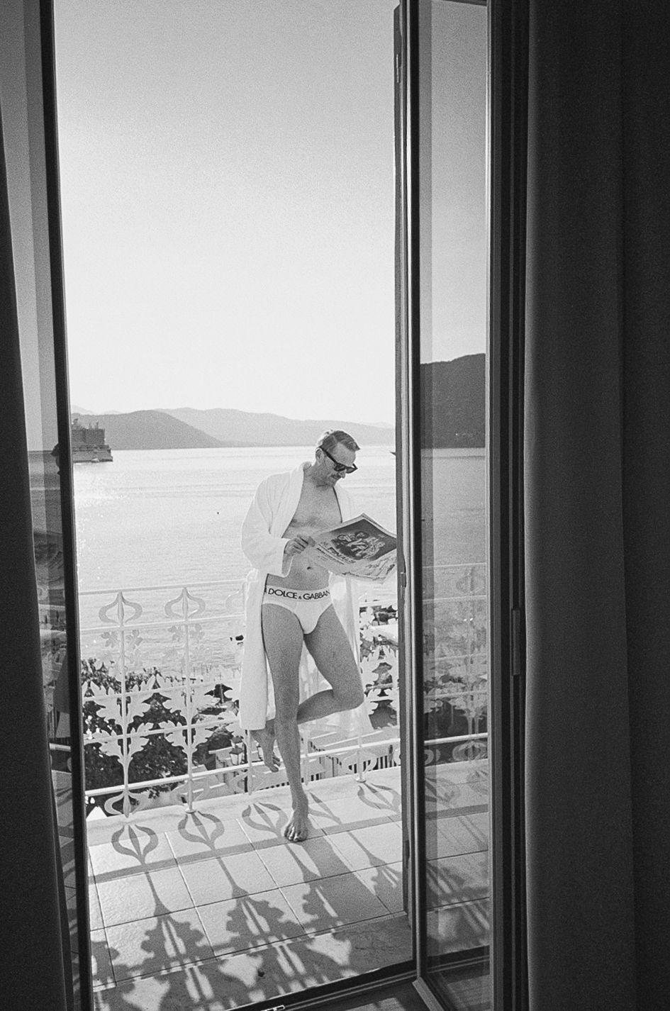 Black and white photo of a man in Dolce & Gabbana underwear reading on a sunlit balcony overlooking a serene lake.