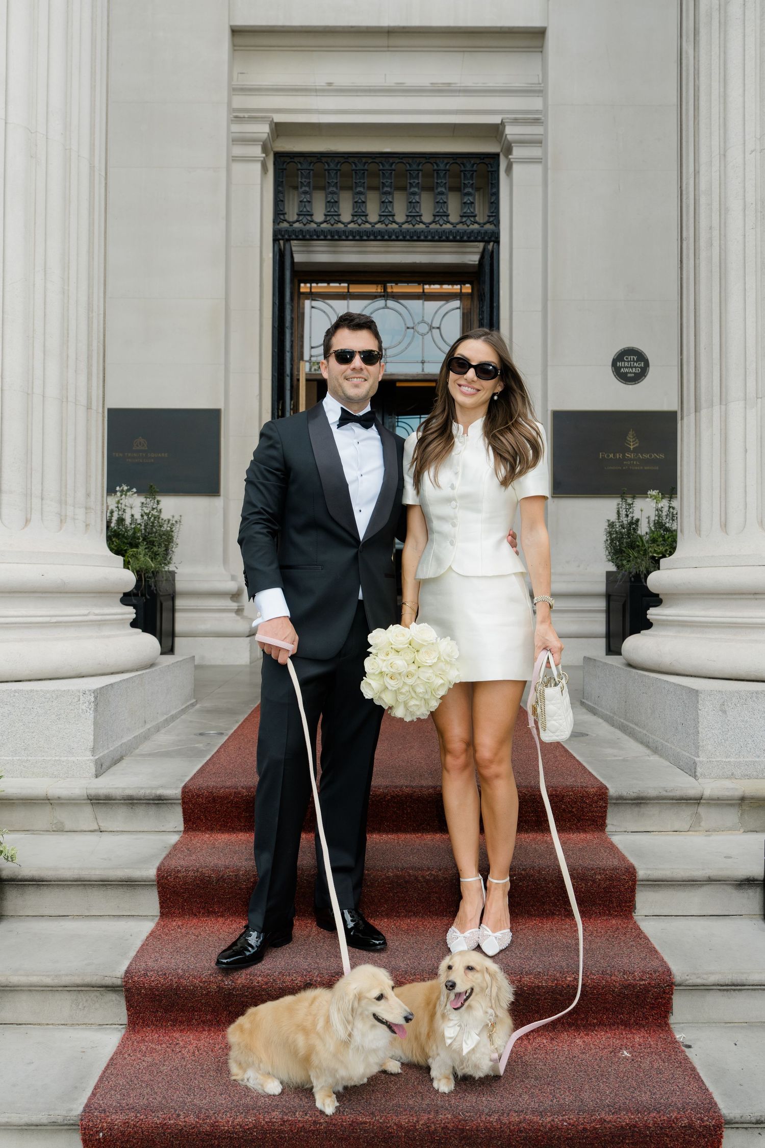 Stylish bride and groom in sunglasses pose with two golden dogs on red carpet hotel steps.