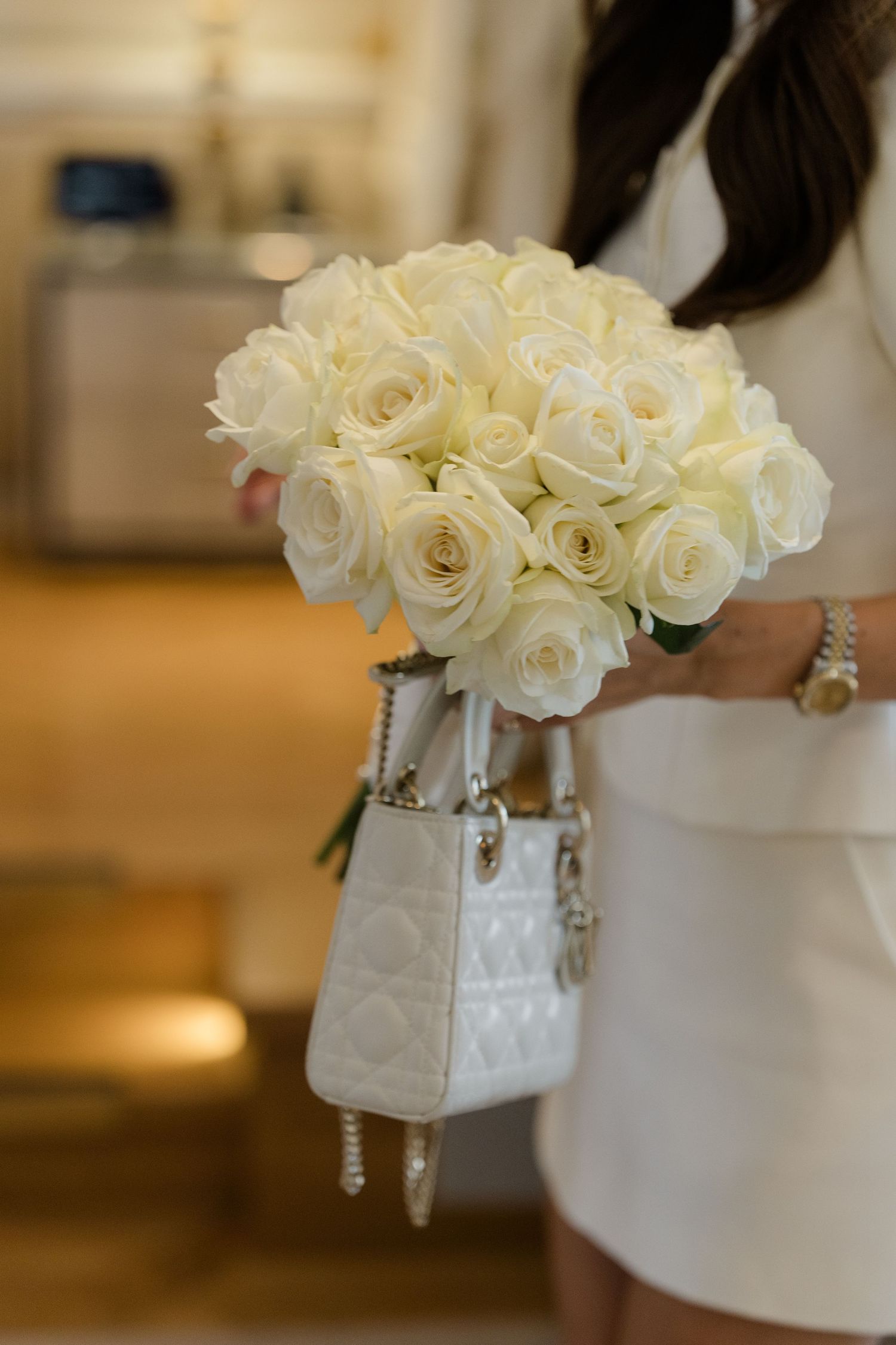 Woman in white dress holding a bouquet of white roses and a quilted white Lady Dior handbag.