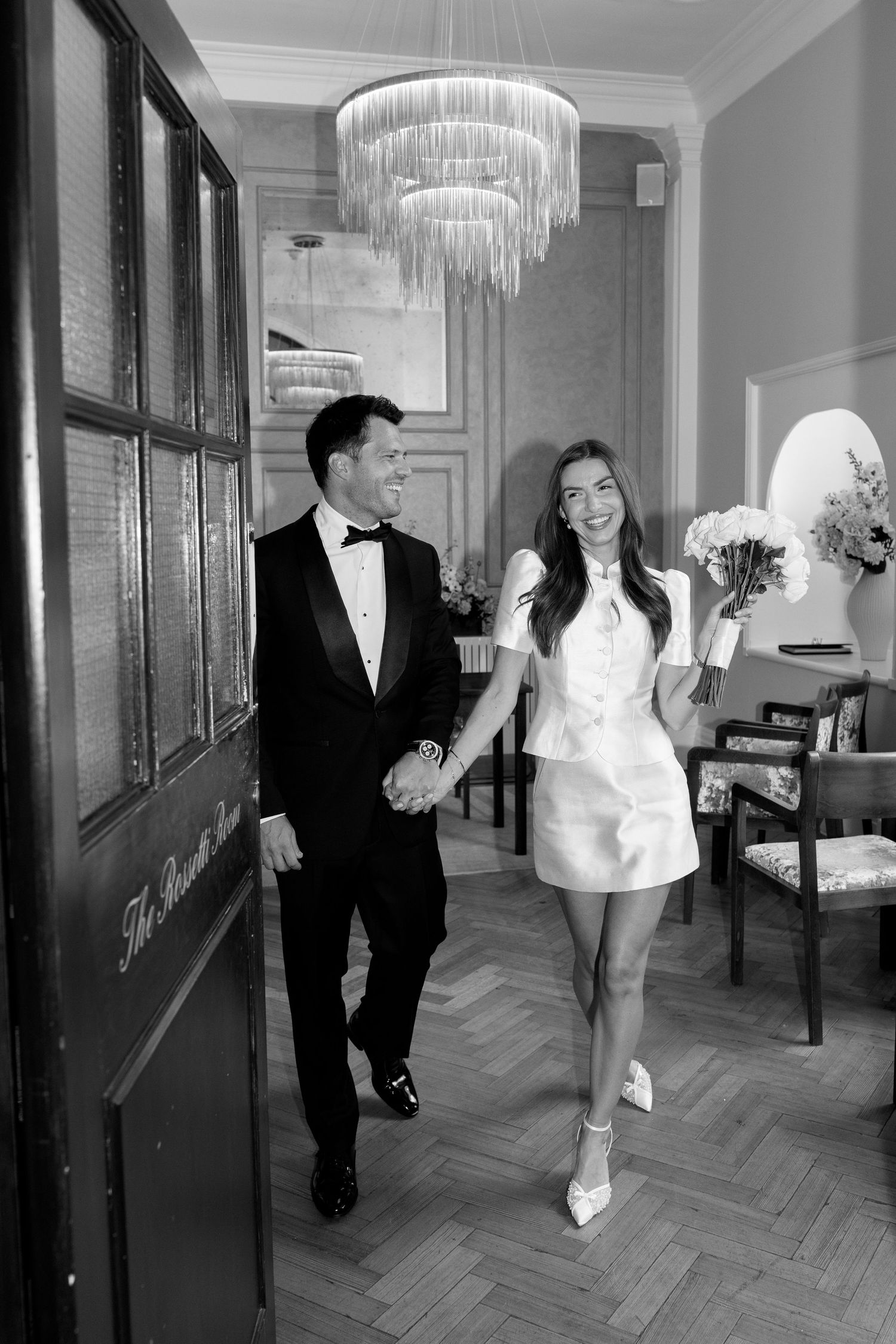 Newlyweds walk hand-in-hand through elegant corridor with chandelier after civil ceremony.