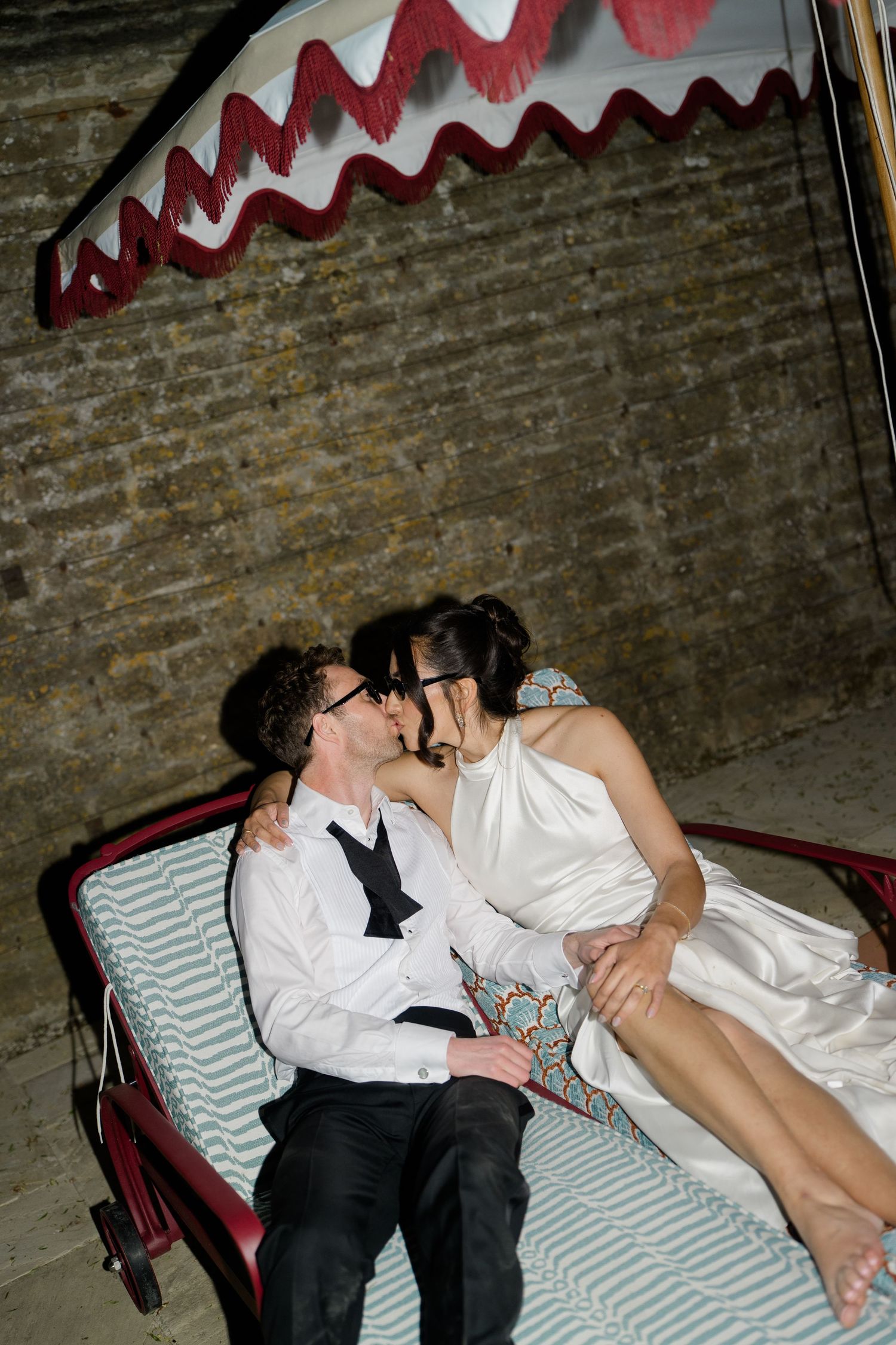 Bride and groom share a playful kiss seated in decorated carriage against rustic brick wall at night.