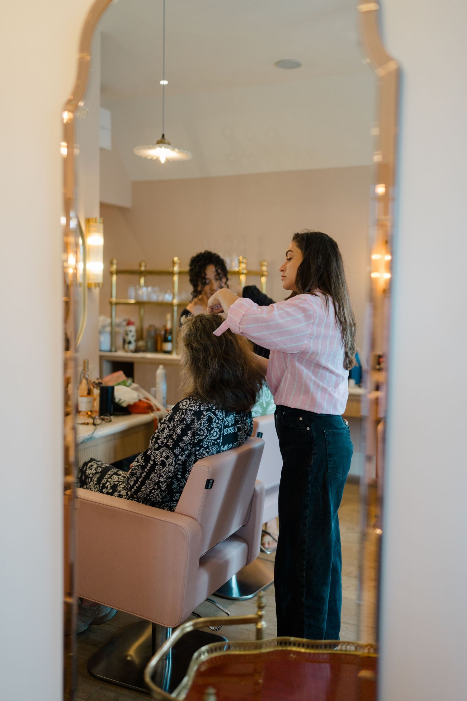 Hair stylist working on client in glamorous salon reflected in ornate gold mirror.