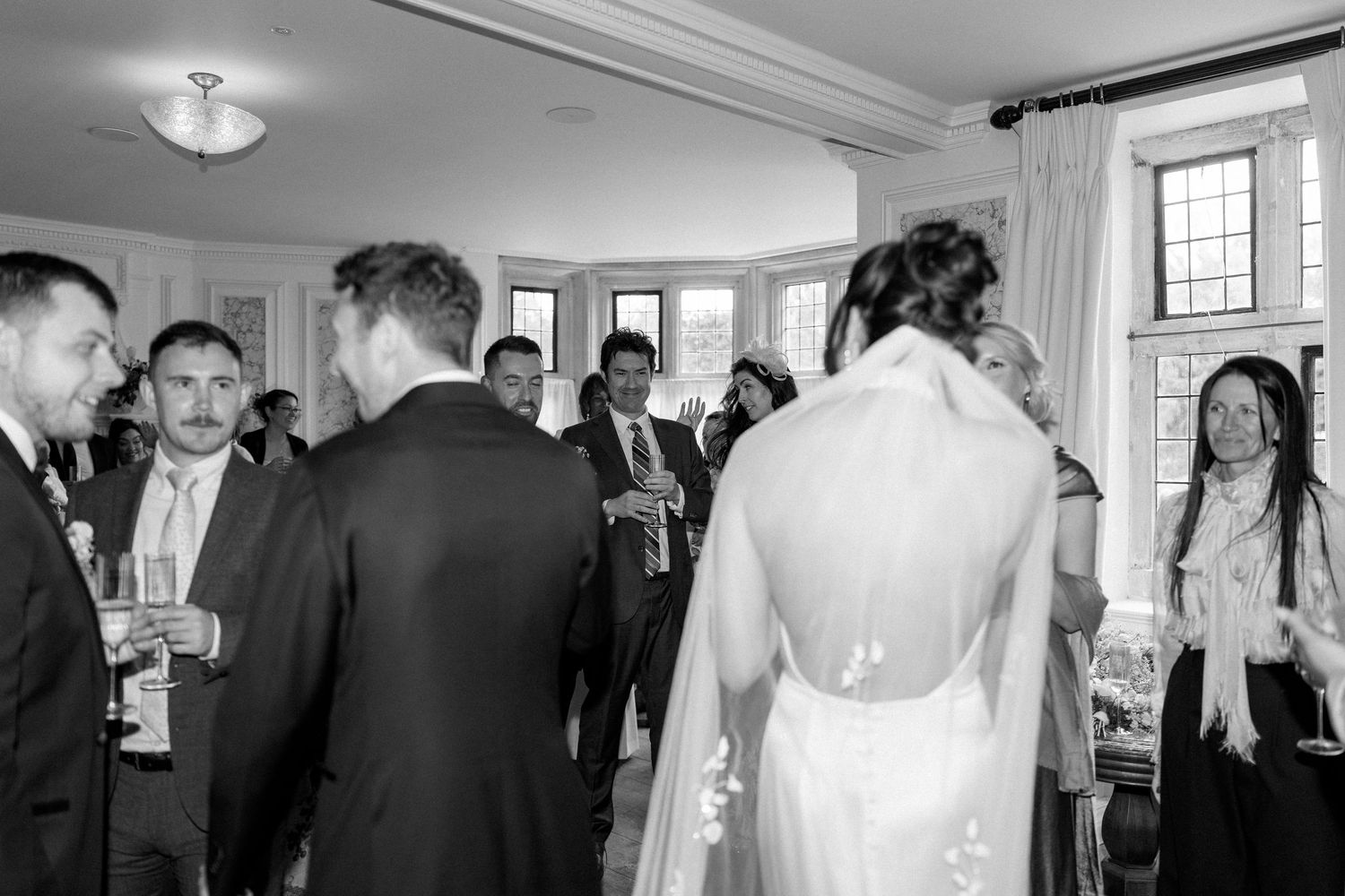 Black and white photo of wedding guests mingling with bride and groom during cocktail hour inside venue.