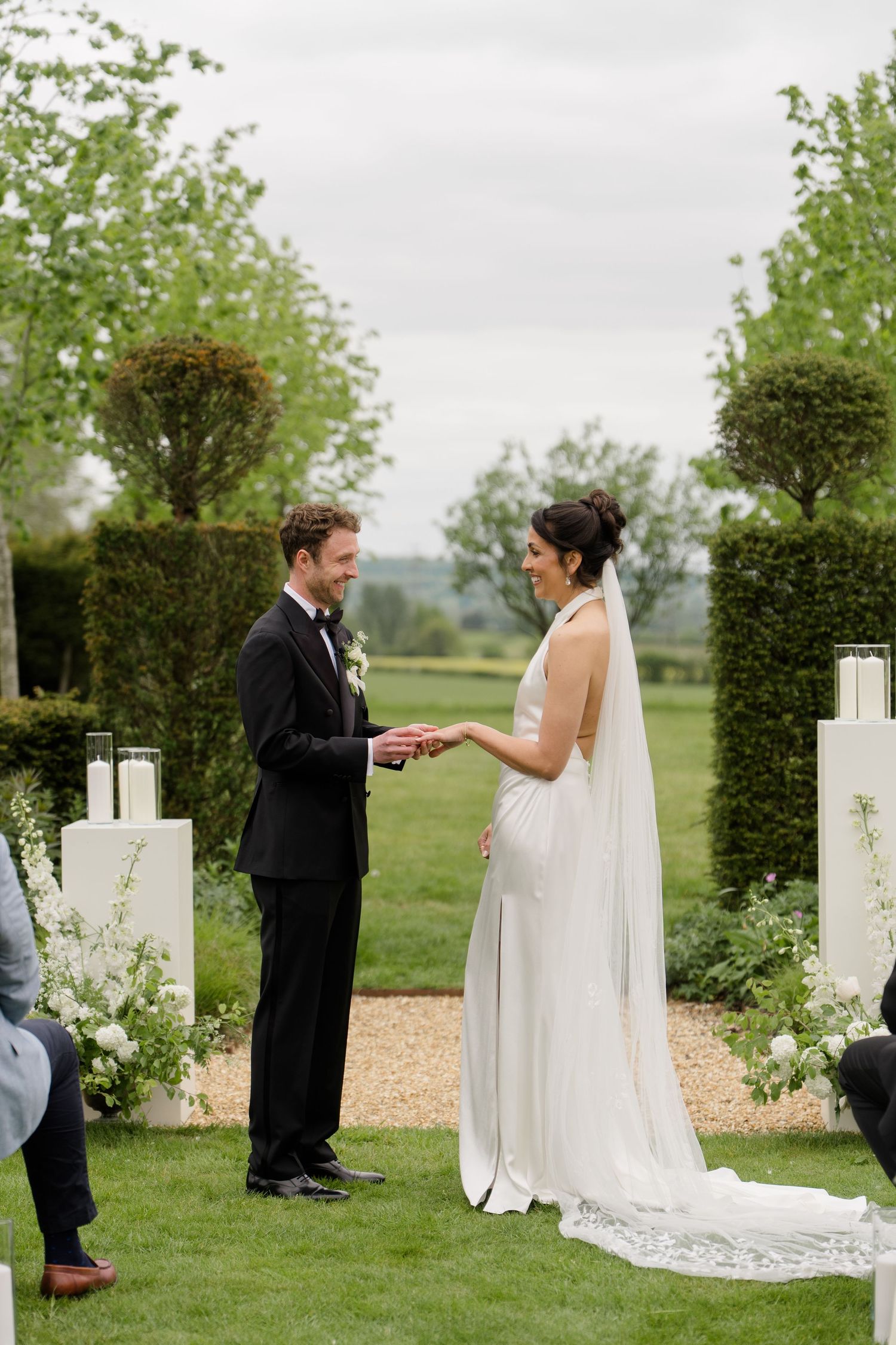 Bride and groom exchange rings during outdoor wedding ceremony surrounded by hedges and white candles.